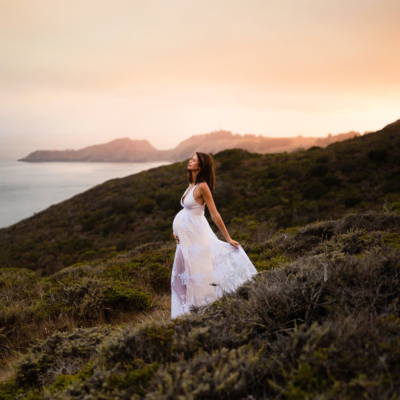 A woman in a flowing white dress stands on a hillside at sunset, with a view of the ocean and distant islands.