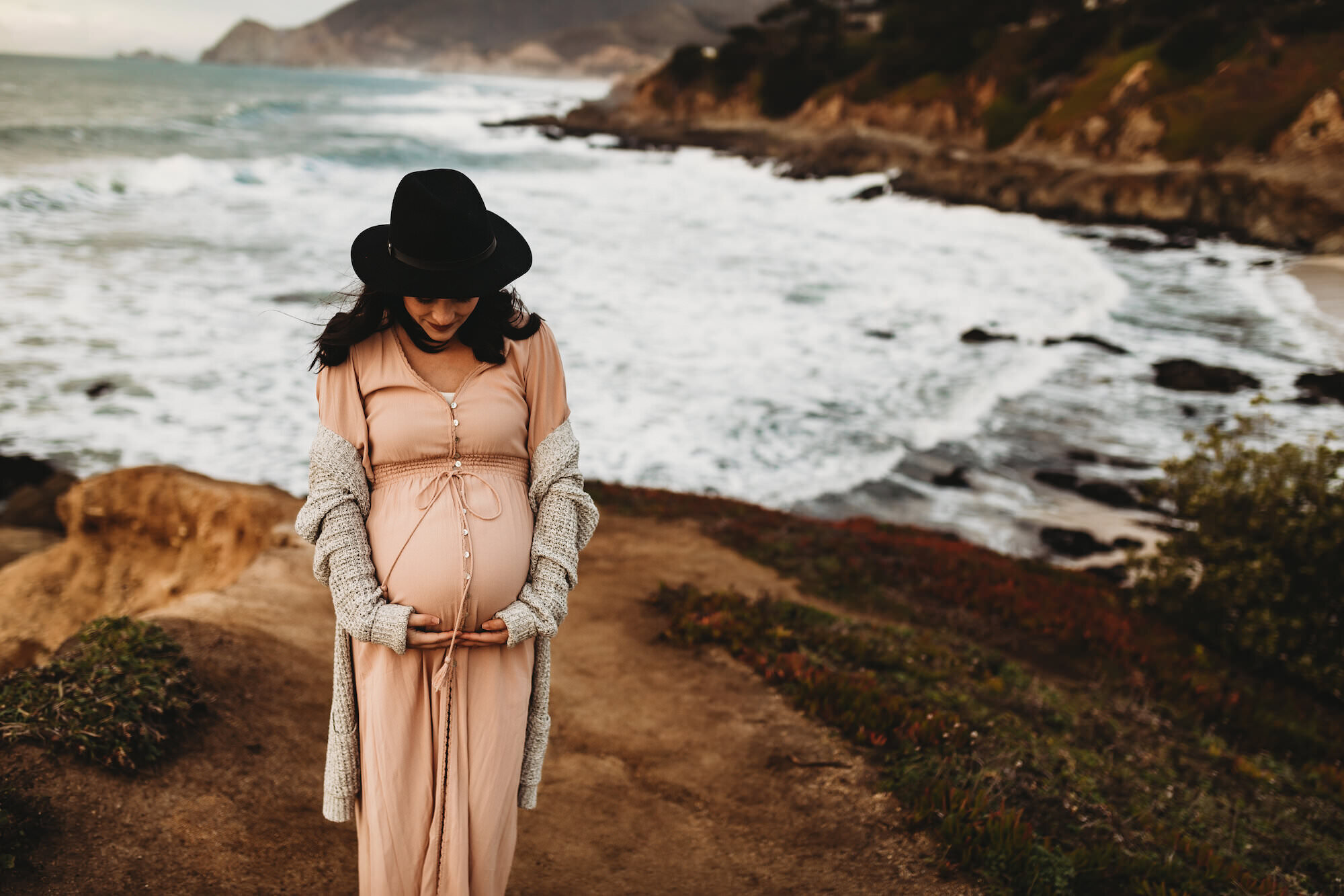 A pregnant woman in a beige dress and a long gray cardigan, wearing a large black hat, standing on a coastal path with cliffs and ocean waves in the background, looking down and holding her belly.