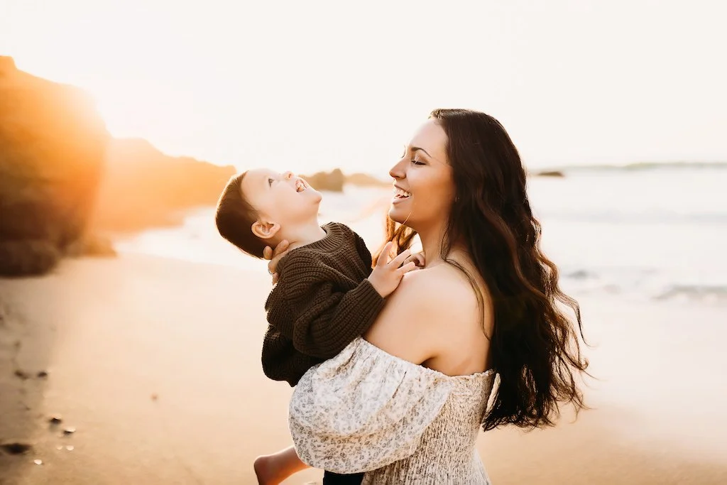 A woman holding a young boy at the beach during sunset, both smiling and looking at each other.