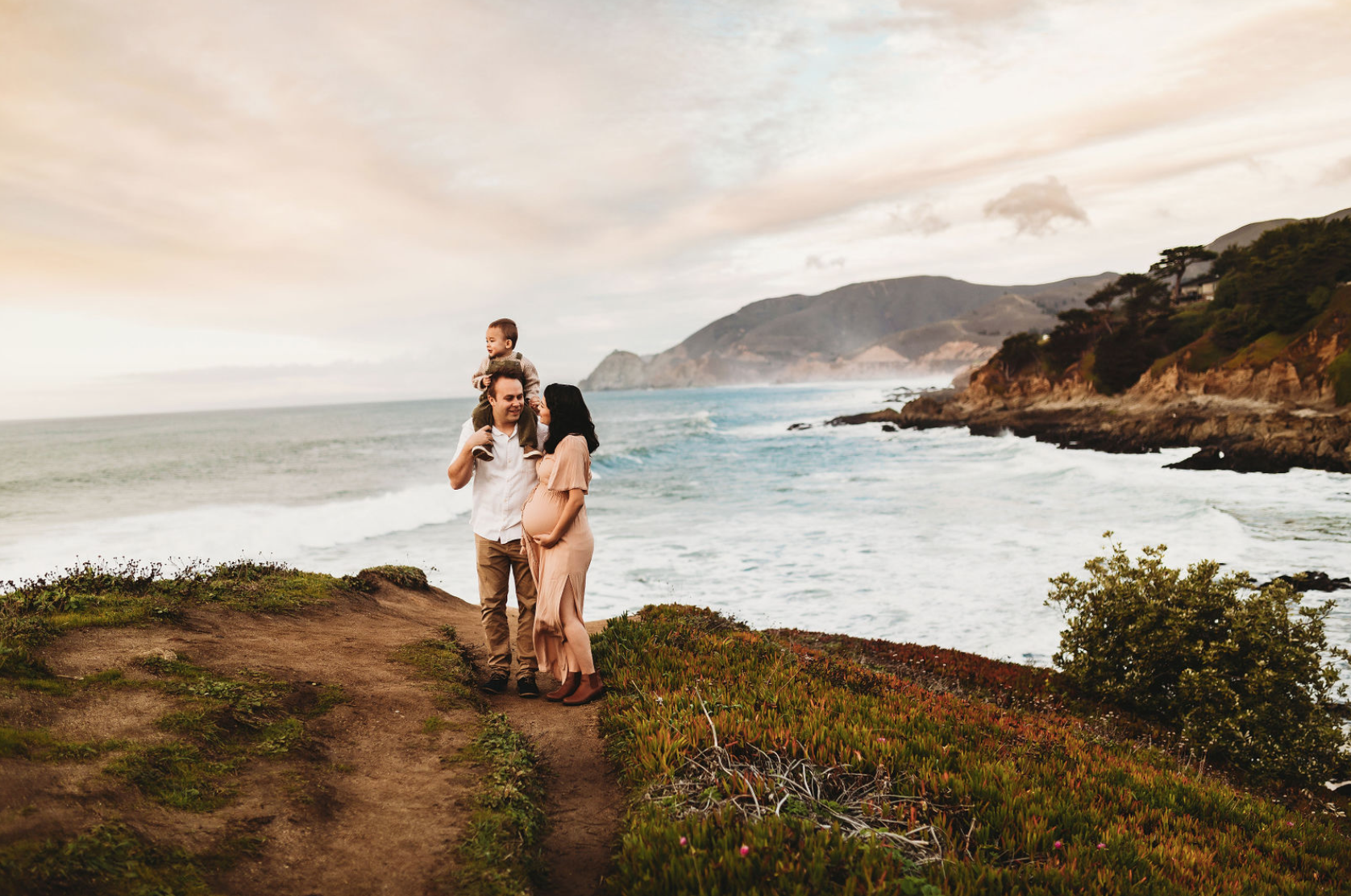 A family of three walking along a coastal path near the ocean with cliffs and hills in the background, during sunset.
