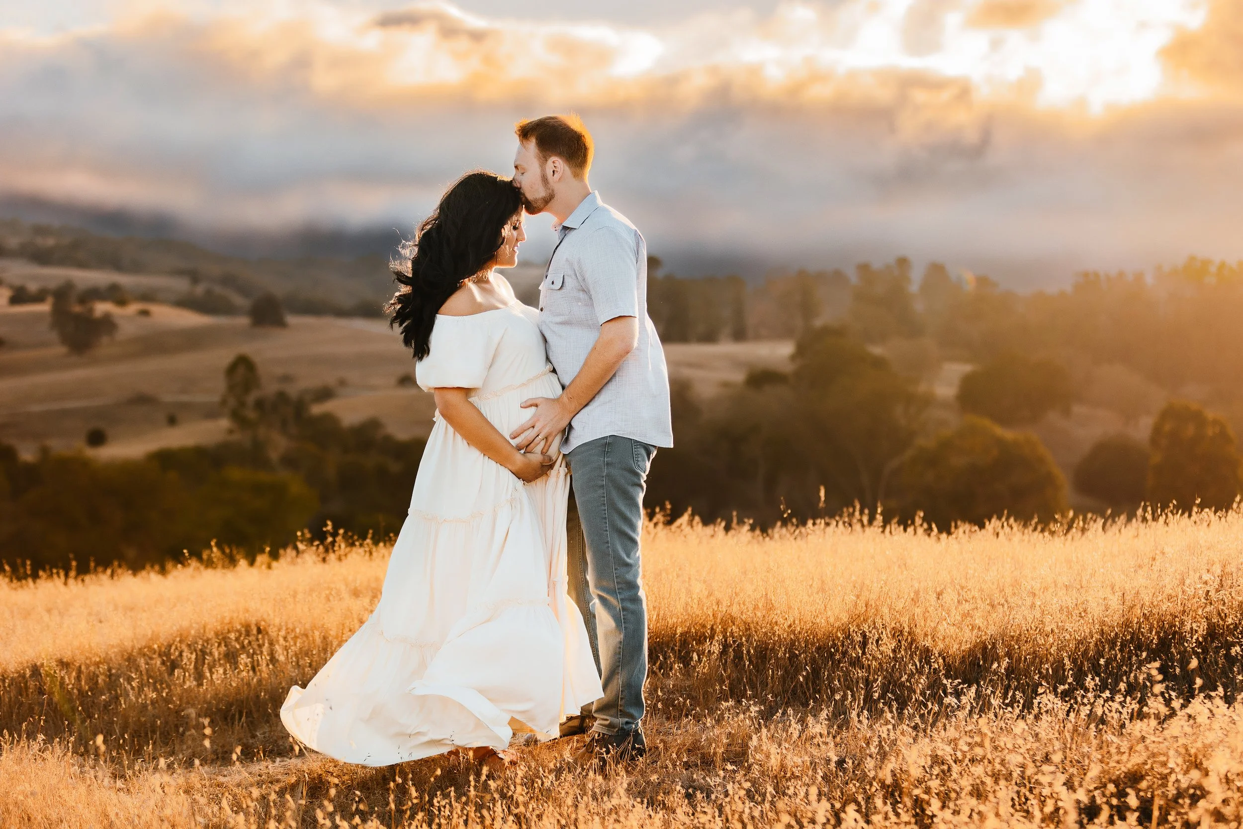 A couple embracing in a field at sunset, with the man kissing the woman's forehead as she leans into him, both standing close together, on a beautiful mountain top in the Bay Area
