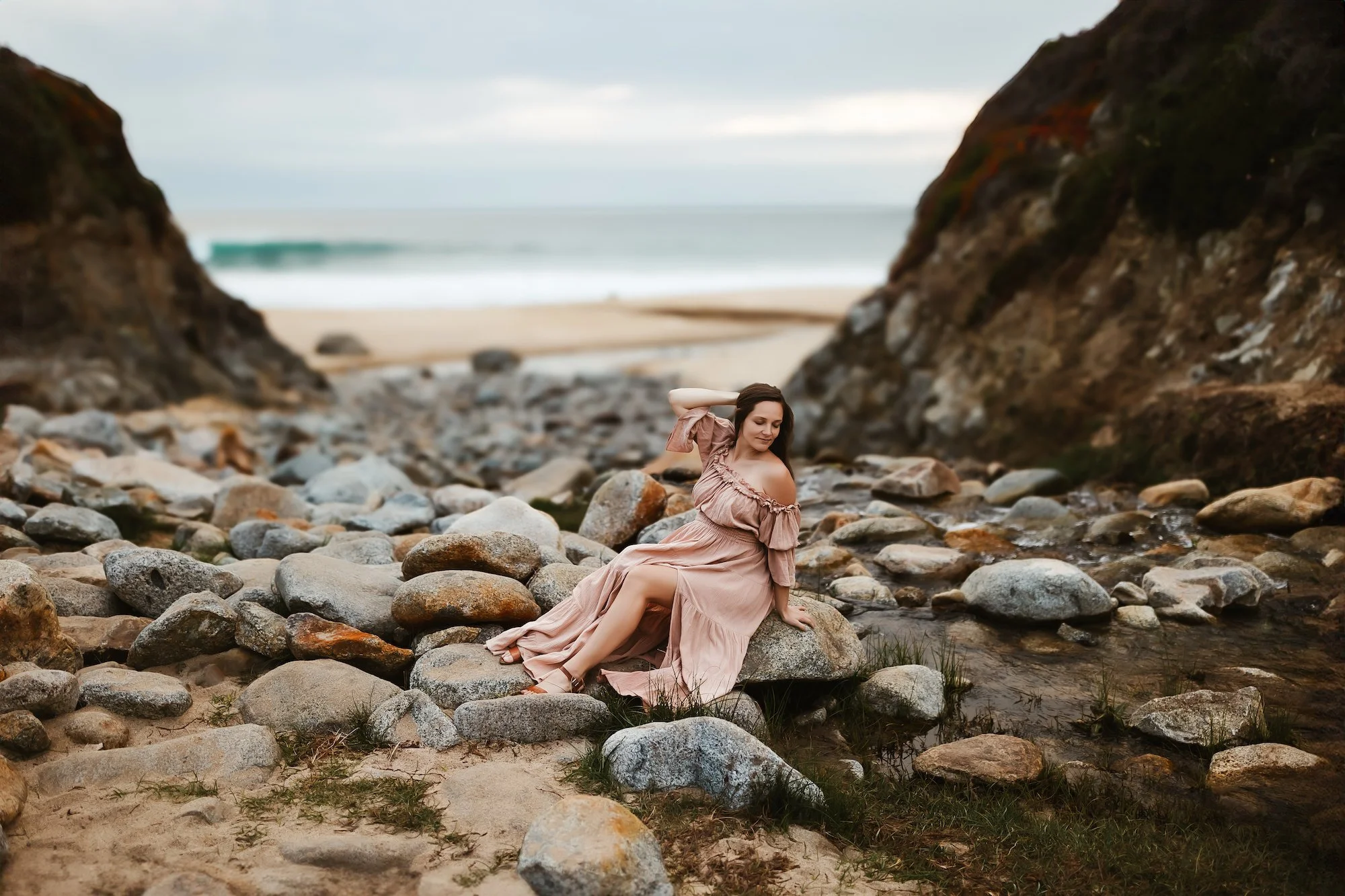 A woman in a pink dress sitting on rocks on a beach near water with cliffs in the background in the Bay Area coast. 