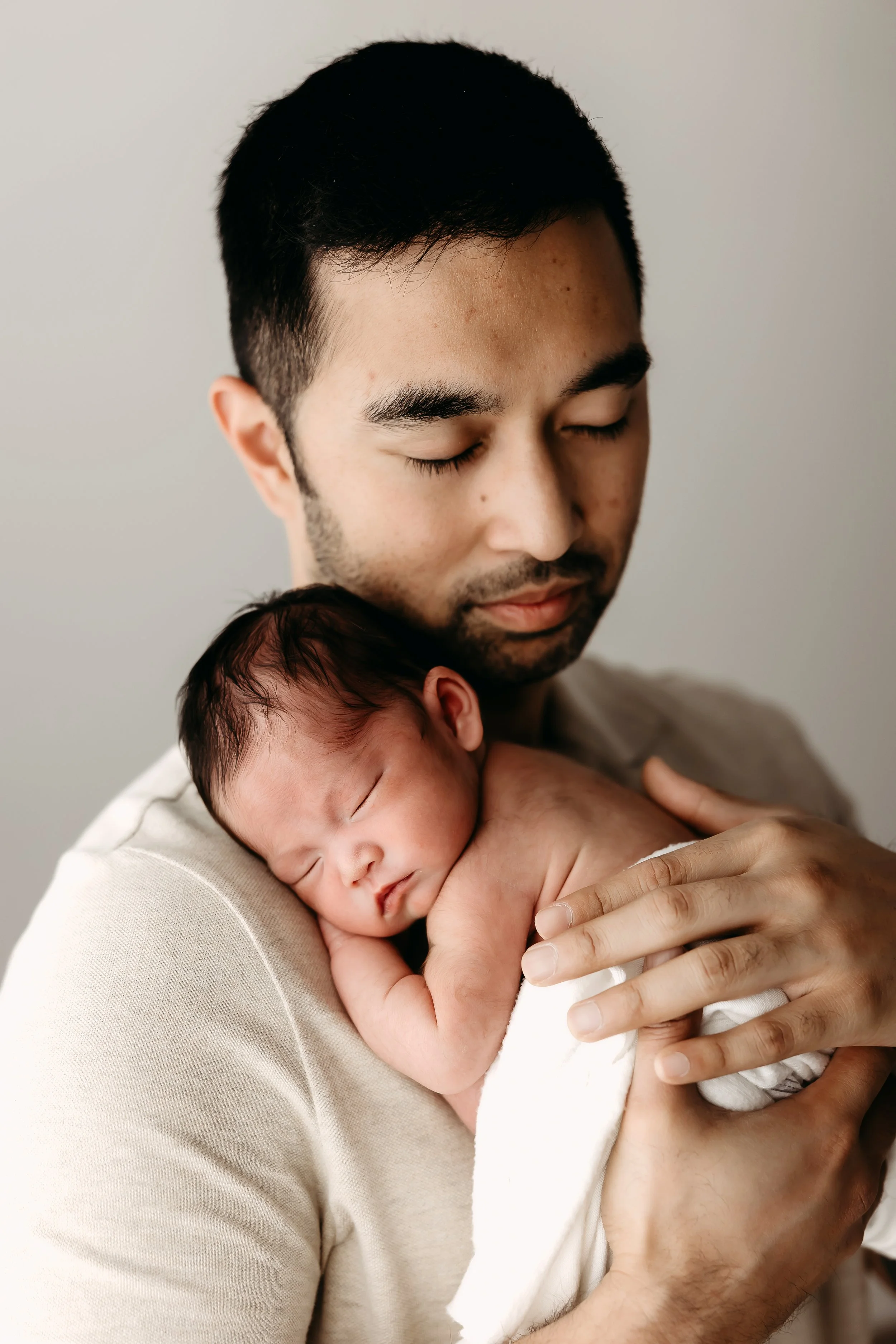 A man with dark hair and a beard cradling a sleeping newborn baby with closed eyes, both with peaceful expressions, against a plain background.