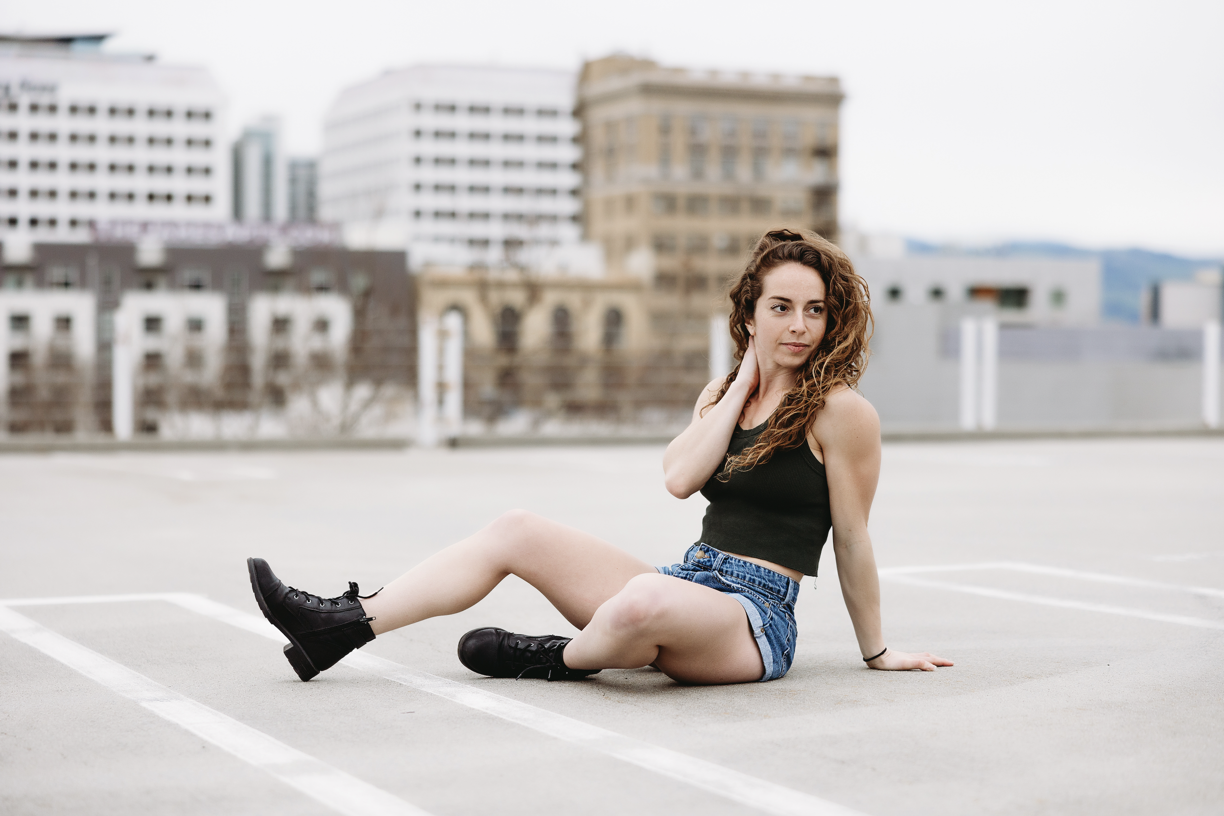 A young woman with curly hair sits on an empty parking lot, with urban buildings in the background.