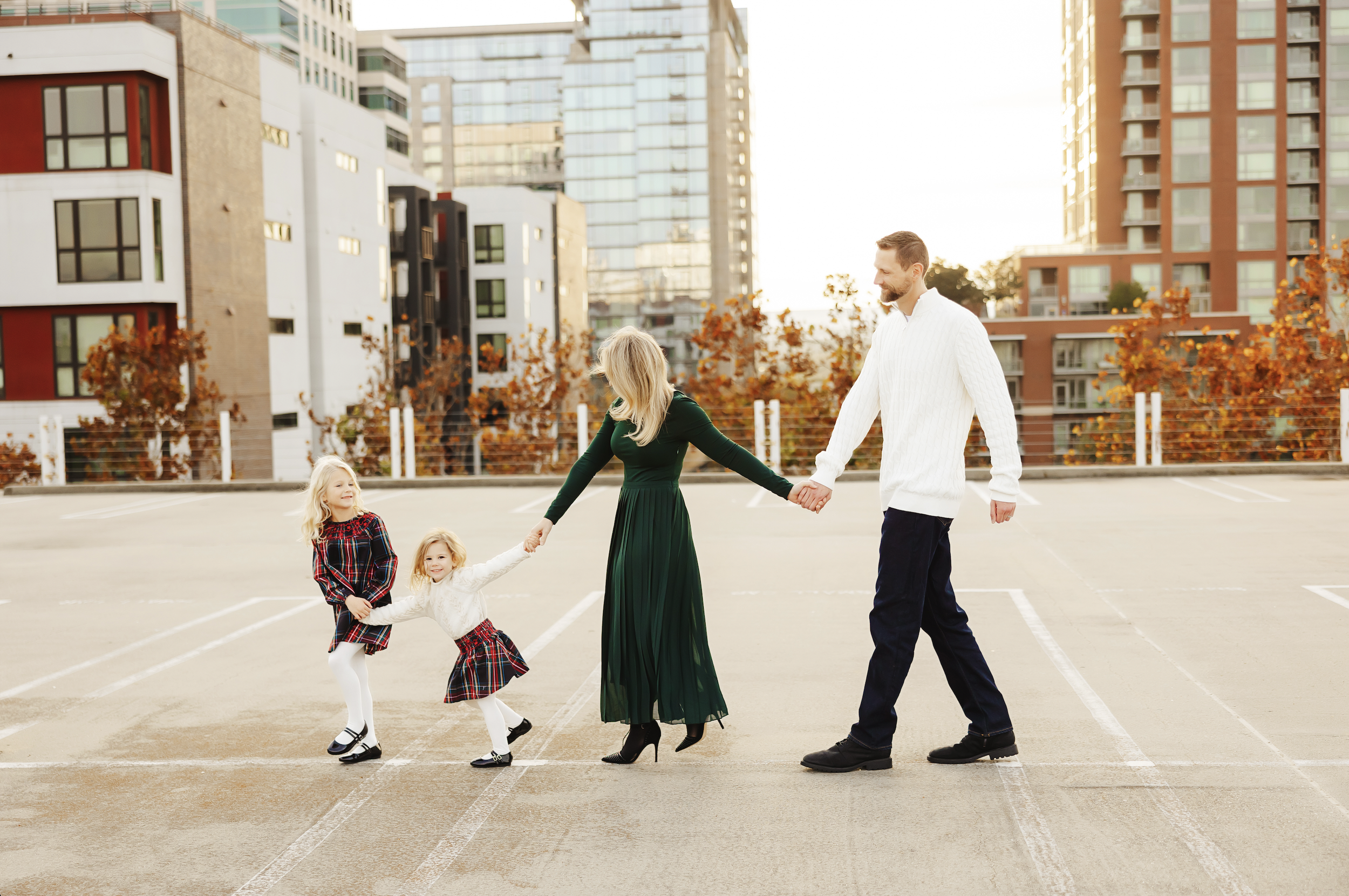 A family of four holding hands and walking in an outdoor parking lot with modern high-rise buildings in the background during the daytime, autumn season.