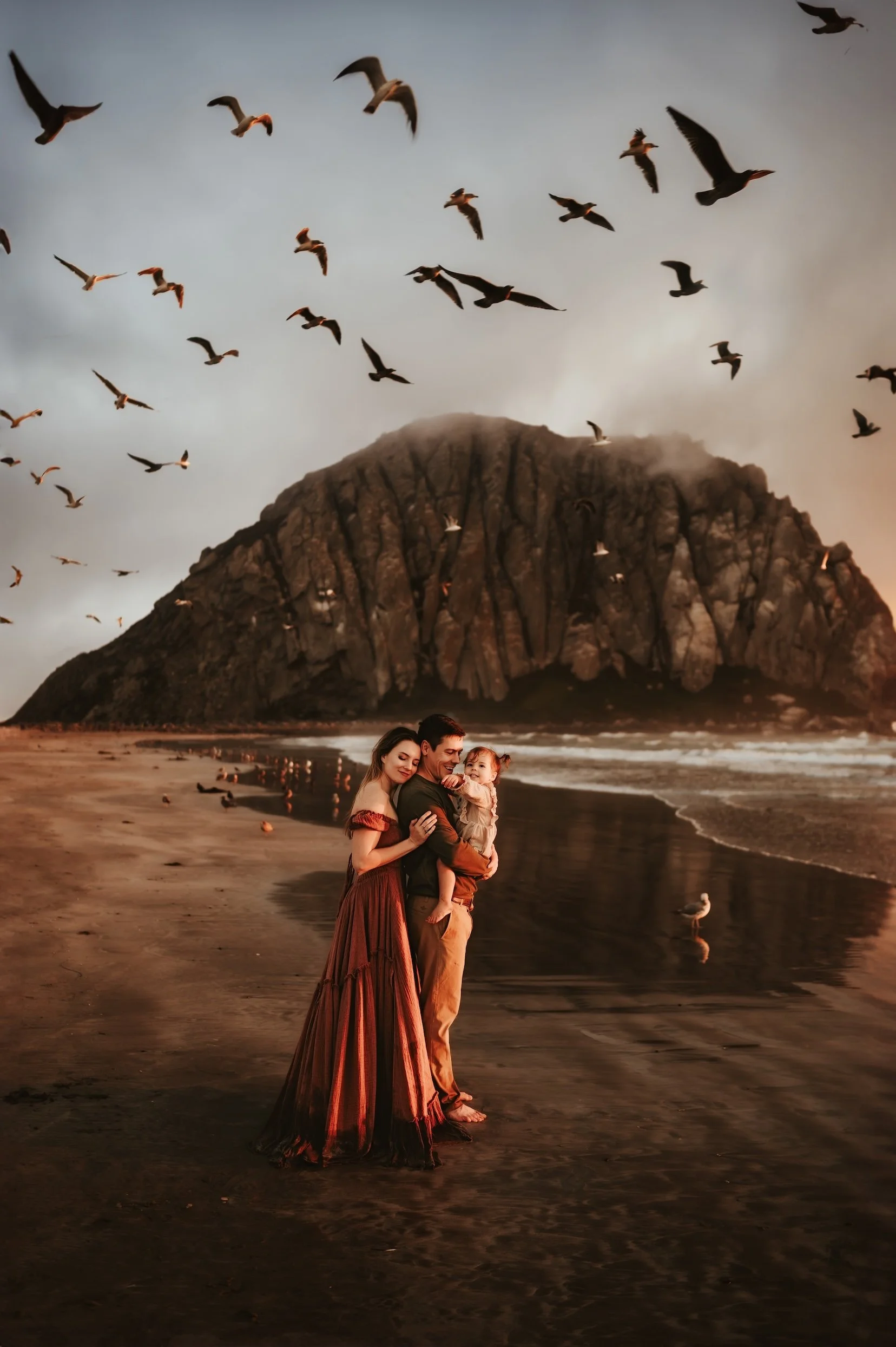 A family of three, a man, woman, and a girl, standing on a beach at sunset with a large rocky mountain in the background. Seagulls fly overhead and some are reflected in the water.
