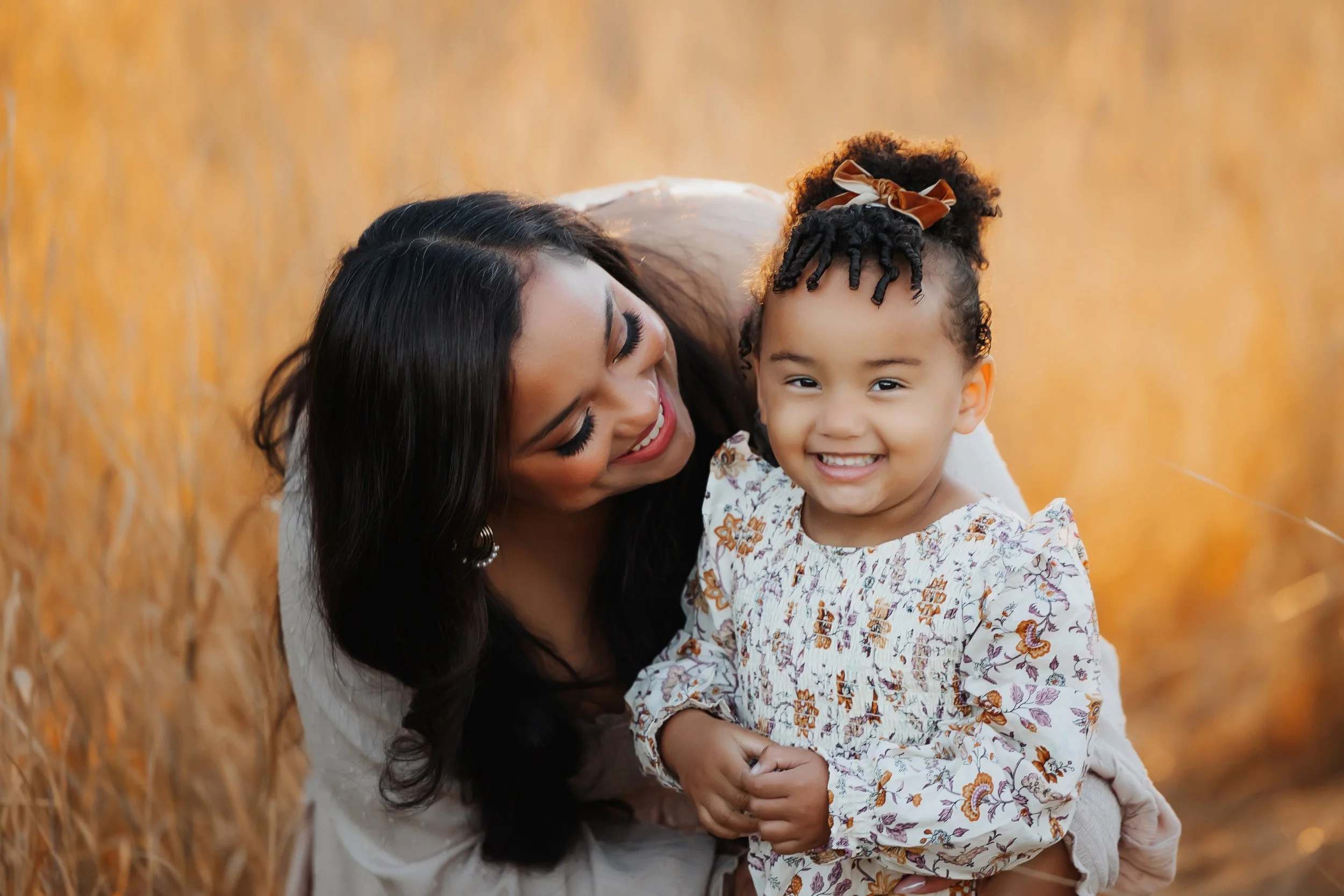 A woman with long dark hair and a girl with curly hair and a bow in her hair smiling and hugging each other in a wheat field during sunset.