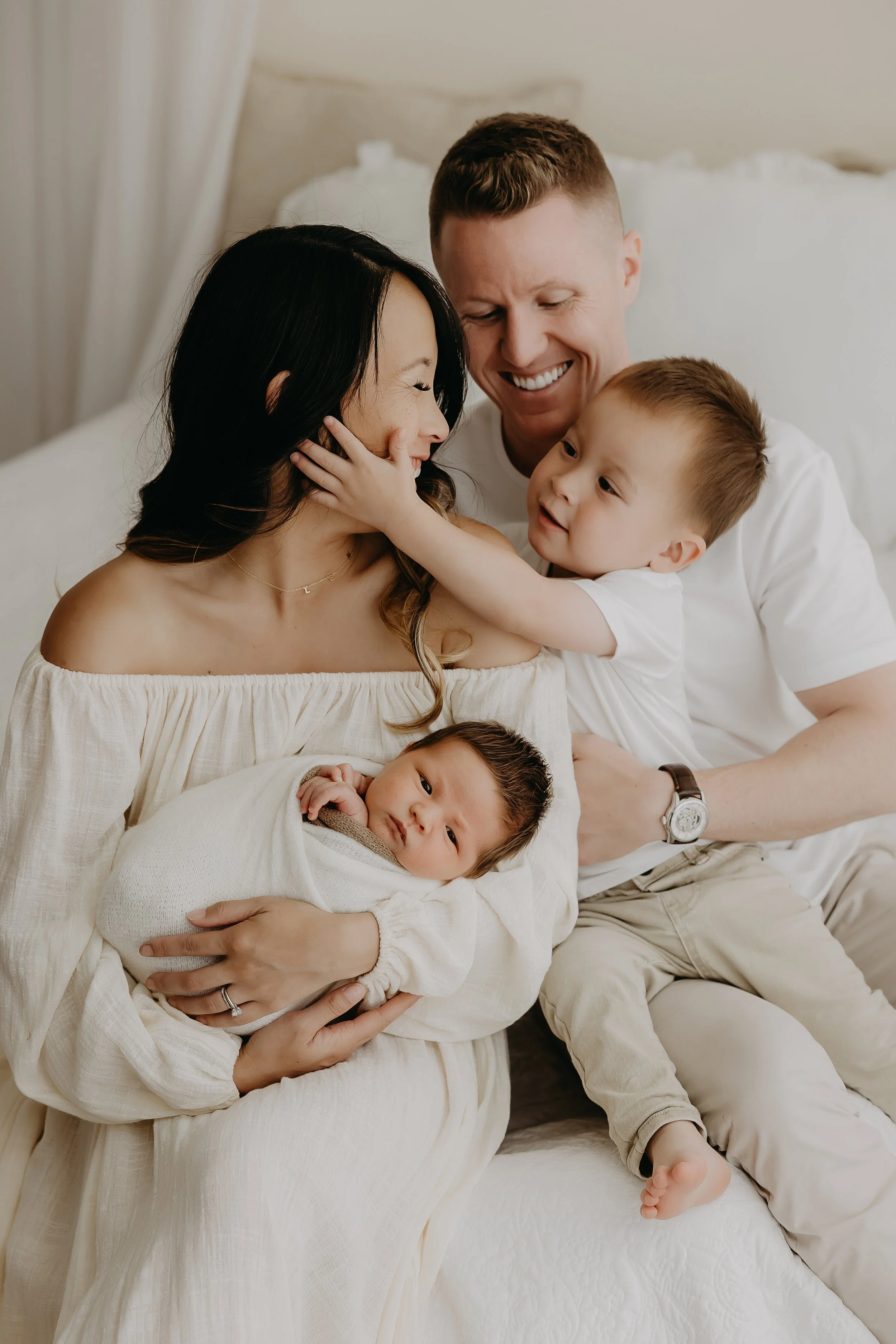 A family of four, including a woman, a man, and two young children, sitting together on a bed and smiling affectionately at each other.