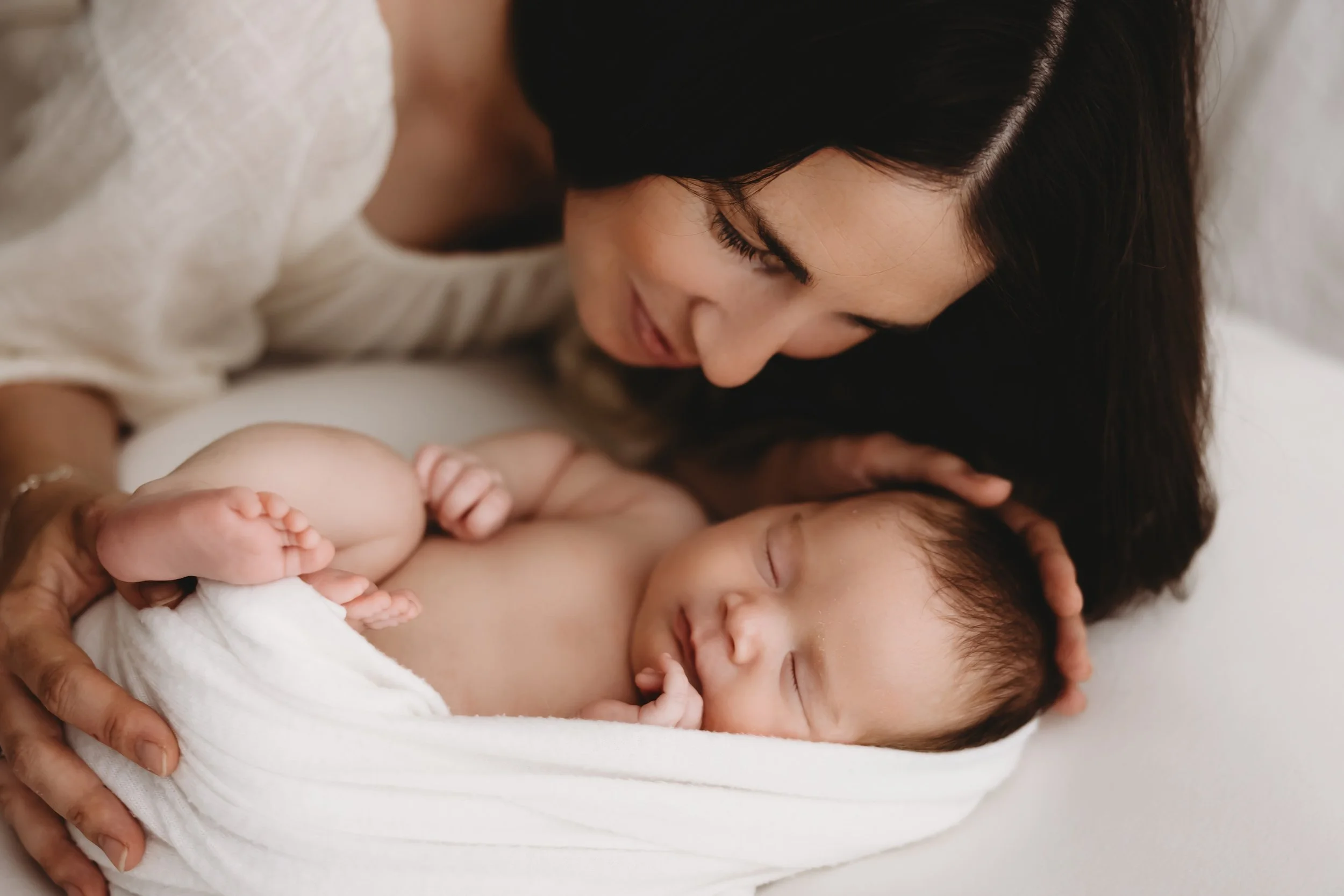 A woman with dark hair lying down and looking at a sleeping baby wrapped in white cloth, holding the baby gently with both hands.