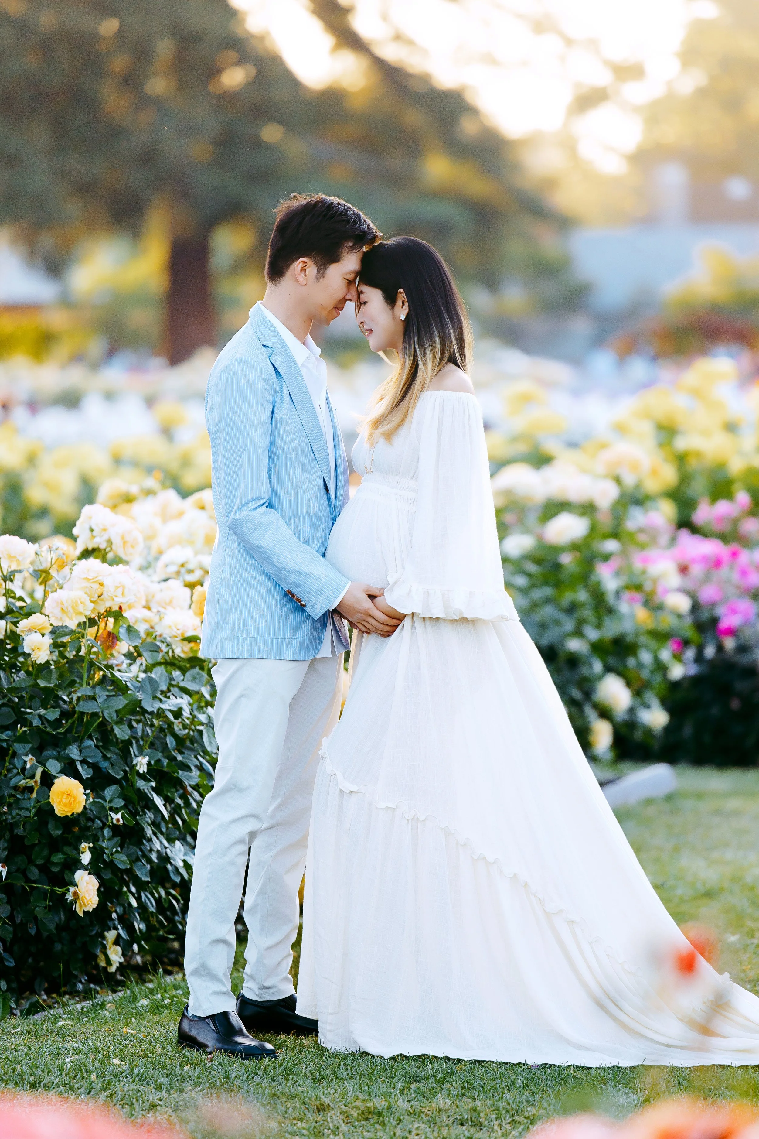 A couple dressed in wedding attire standing in a garden with yellow and pink roses, embracing with their foreheads touching, during sunset.