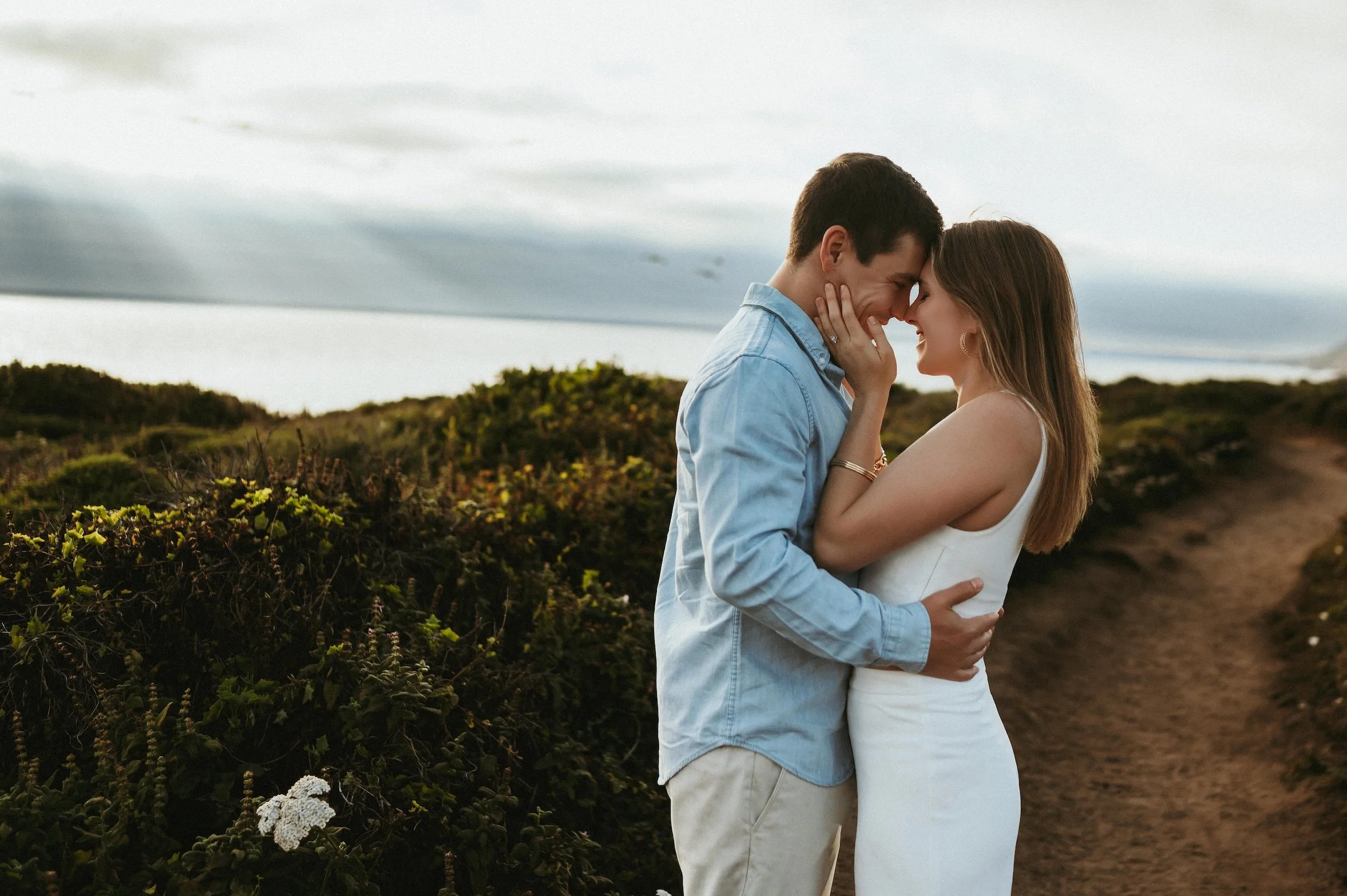 A couple is standing very close together outdoors on a cloudy day, touching foreheads and holding each other affectionately amid green bushes and wildflowers.