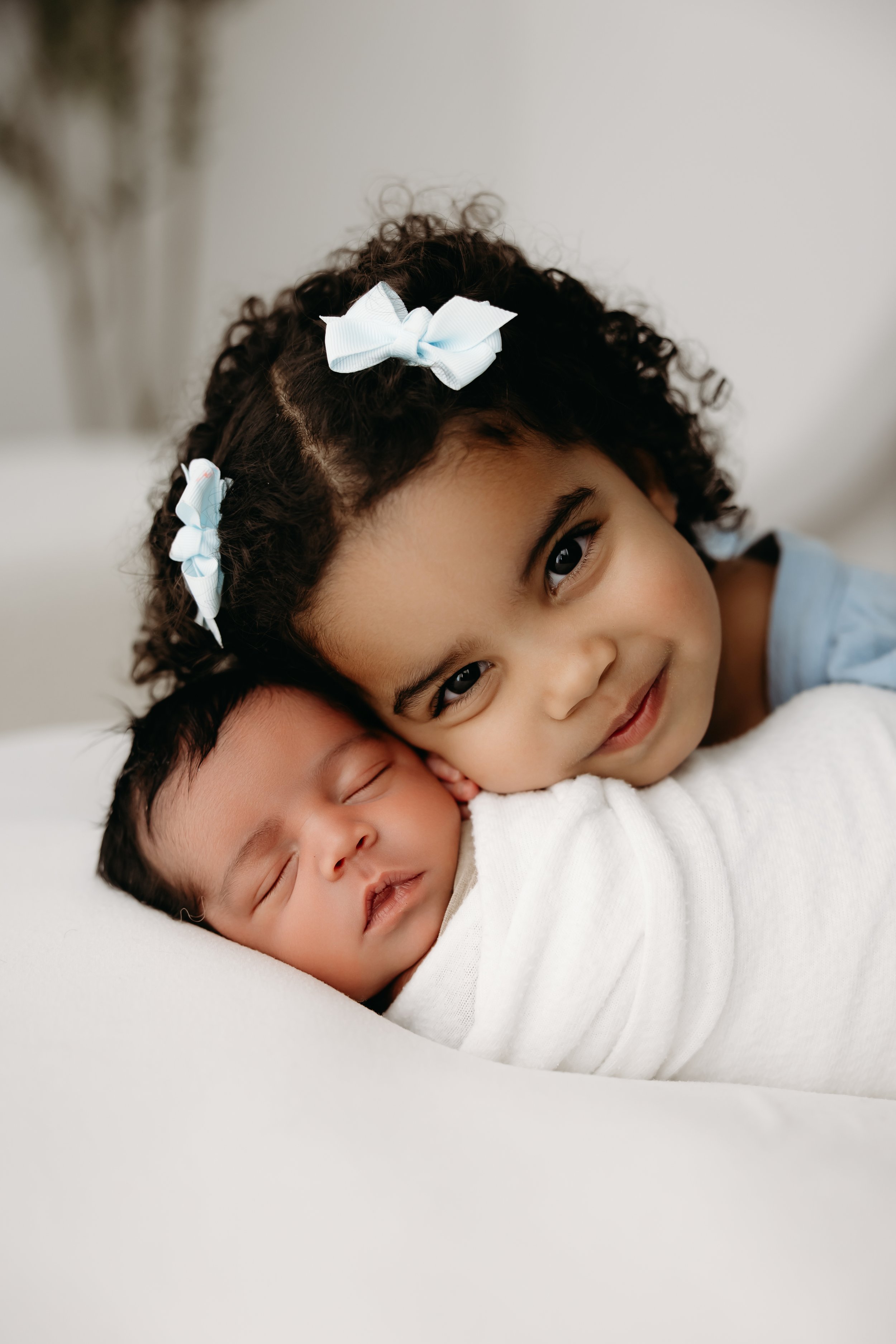 A young girl with curly hair and bows, gently hugging a sleeping baby with closed eyes, both resting on a white surface.