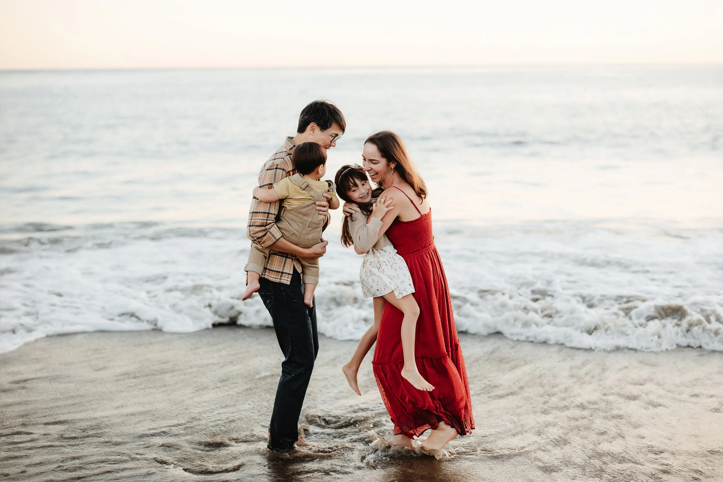 A family with two children enjoying their time at the beach as the mother and father hold their kids close and walk in the shallow water, with gentle waves and a bright sky in the background.