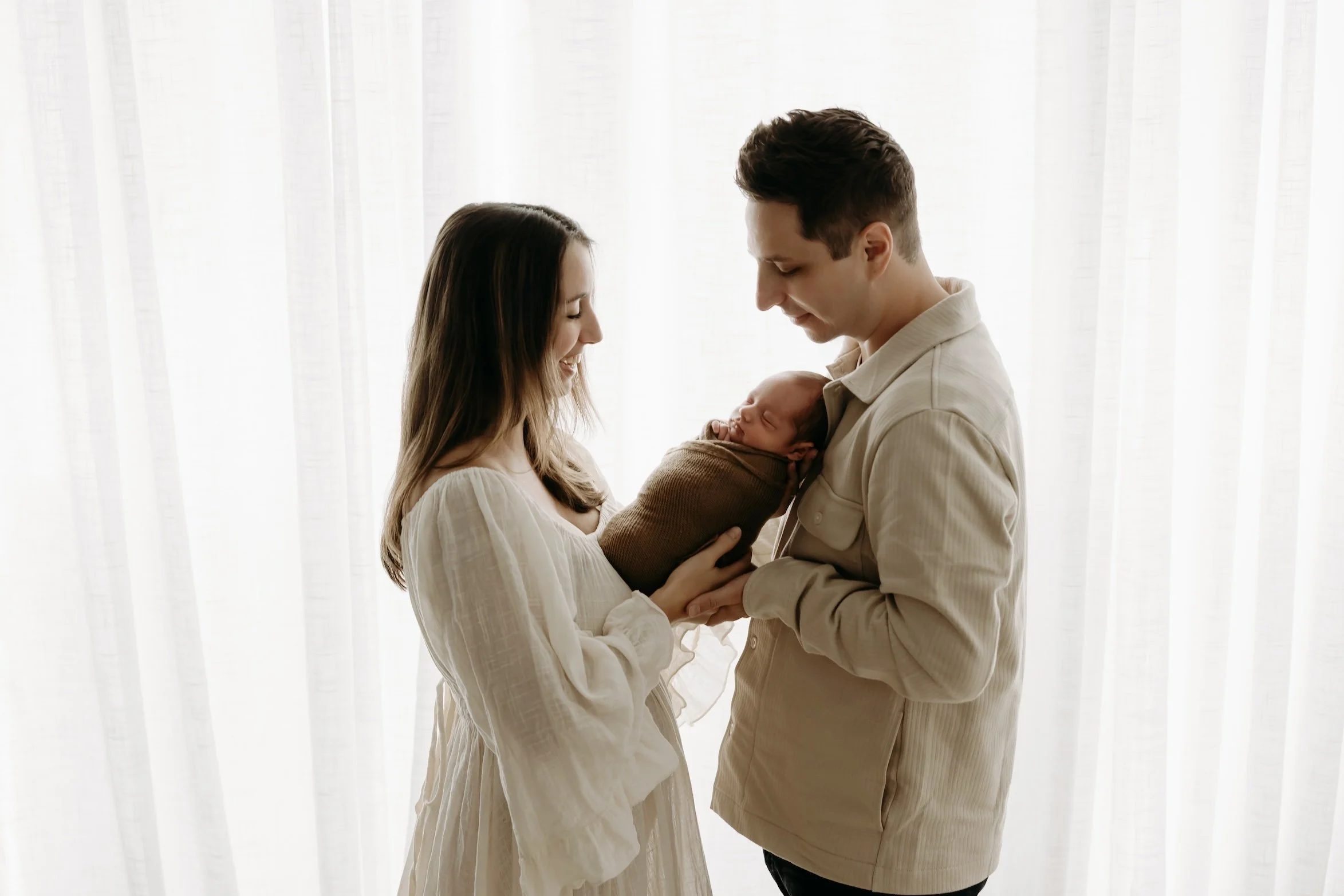 A family of three, a woman, a man, and a newborn baby, stands against a white curtain background. The woman and man are smiling and looking at the baby, who is wrapped in a blanket and held by the woman.