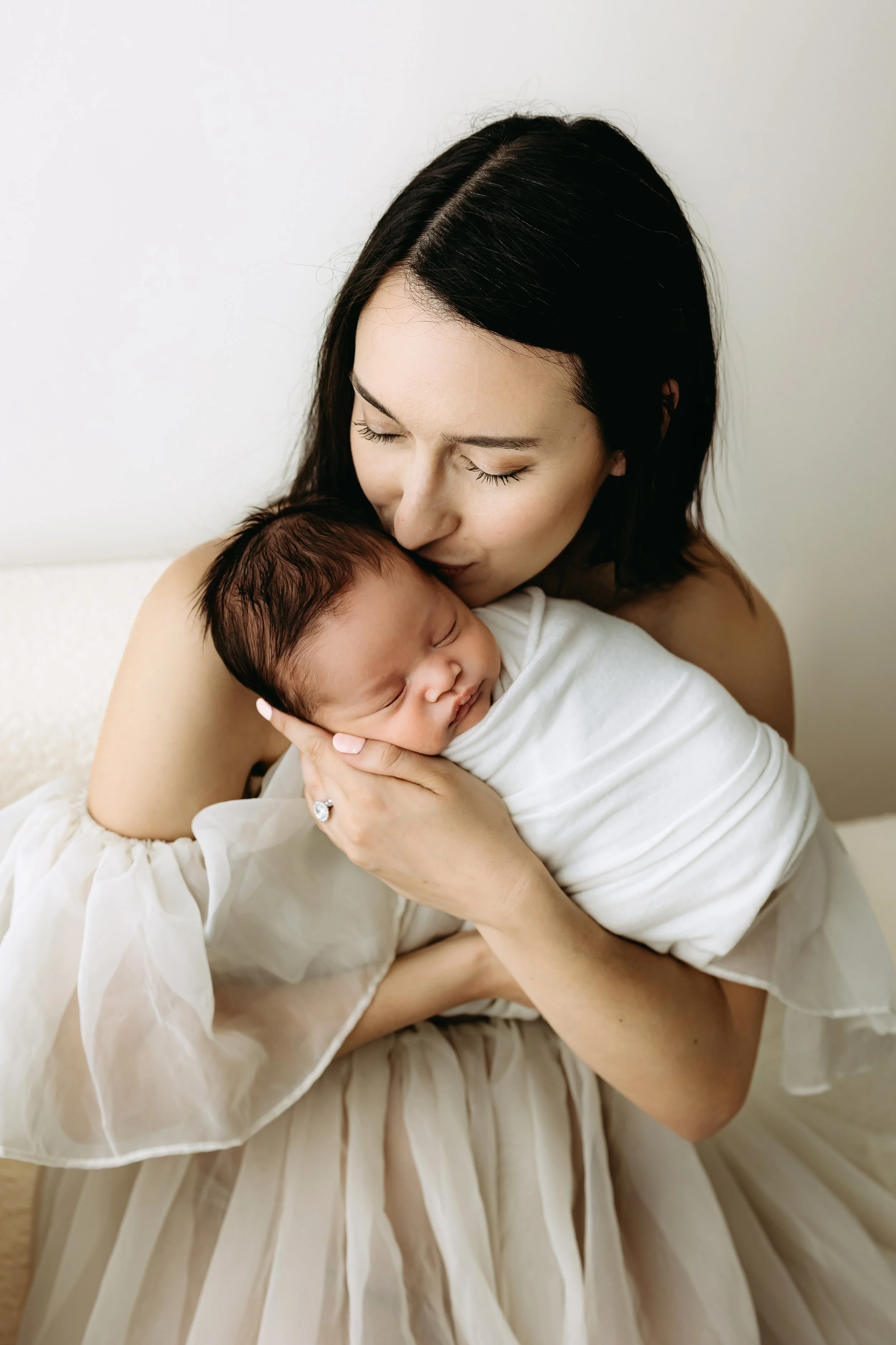 A woman with dark hair holding a sleeping baby wrapped in a white blanket.