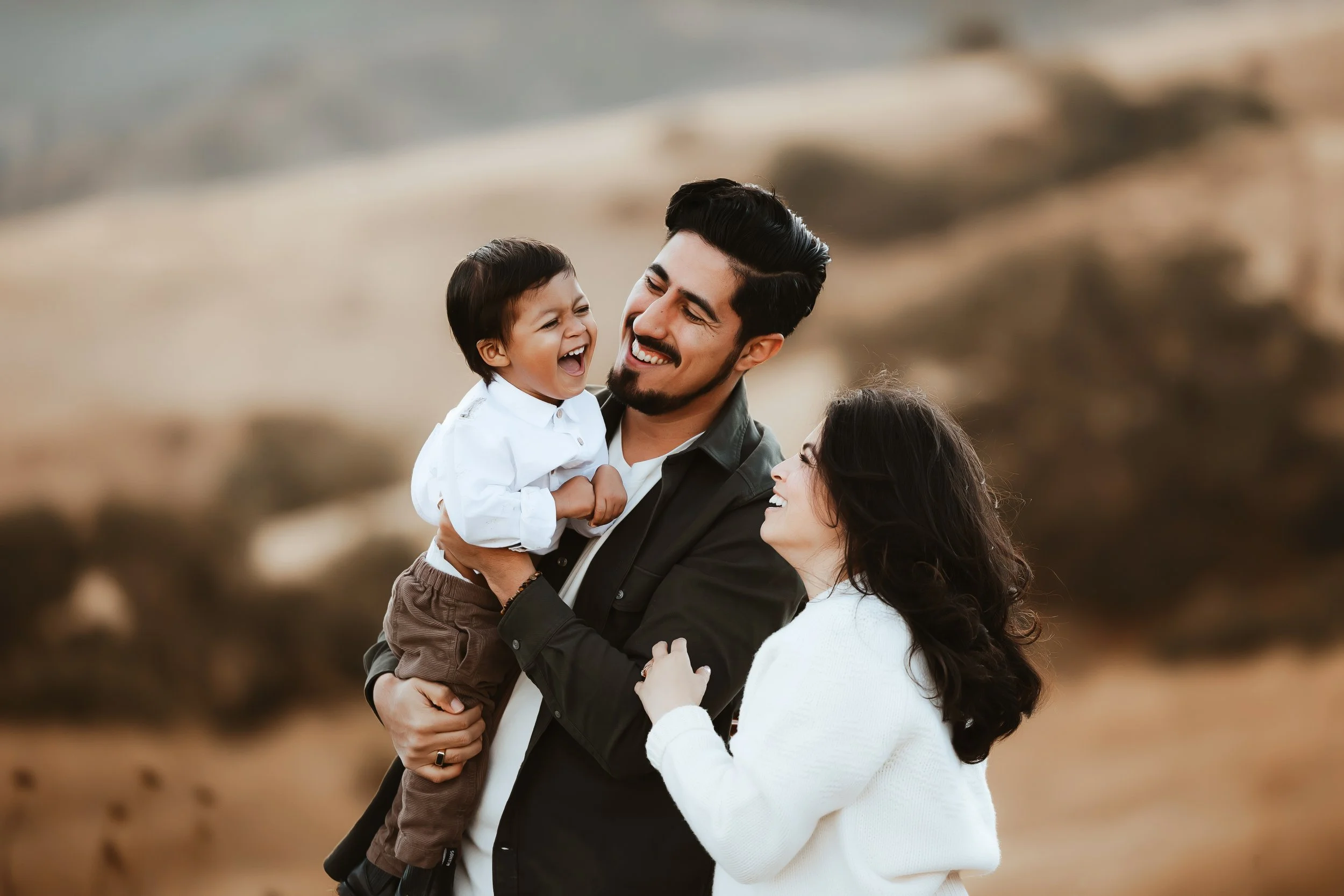 A joyful family outdoors: a man holding a young boy and a woman looking at the boy, all smiling and laughing.
