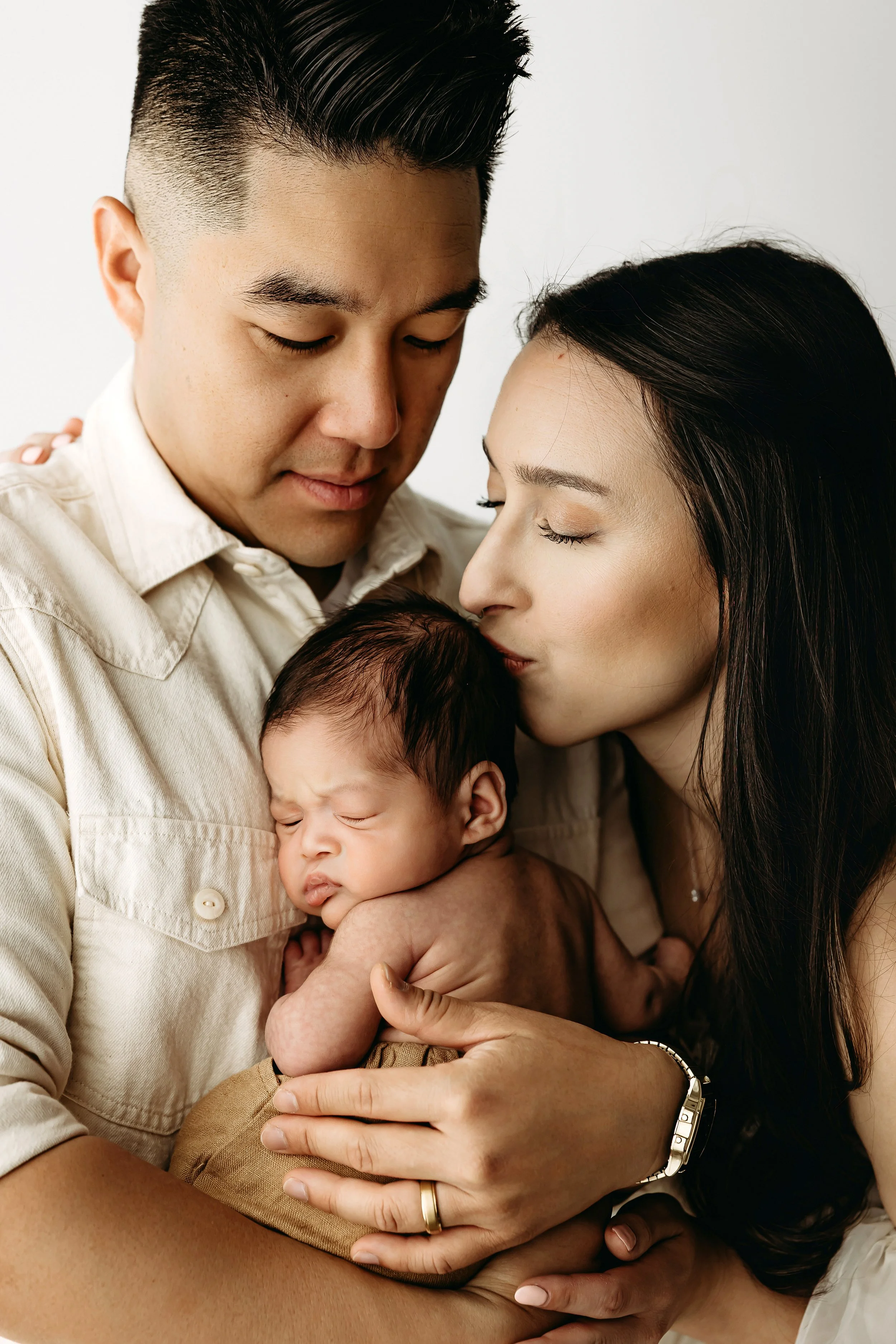 A multicultural family of three, a man, a woman, and a baby, sharing a close, intimate moment with their eyes closed, embracing each other tenderly.