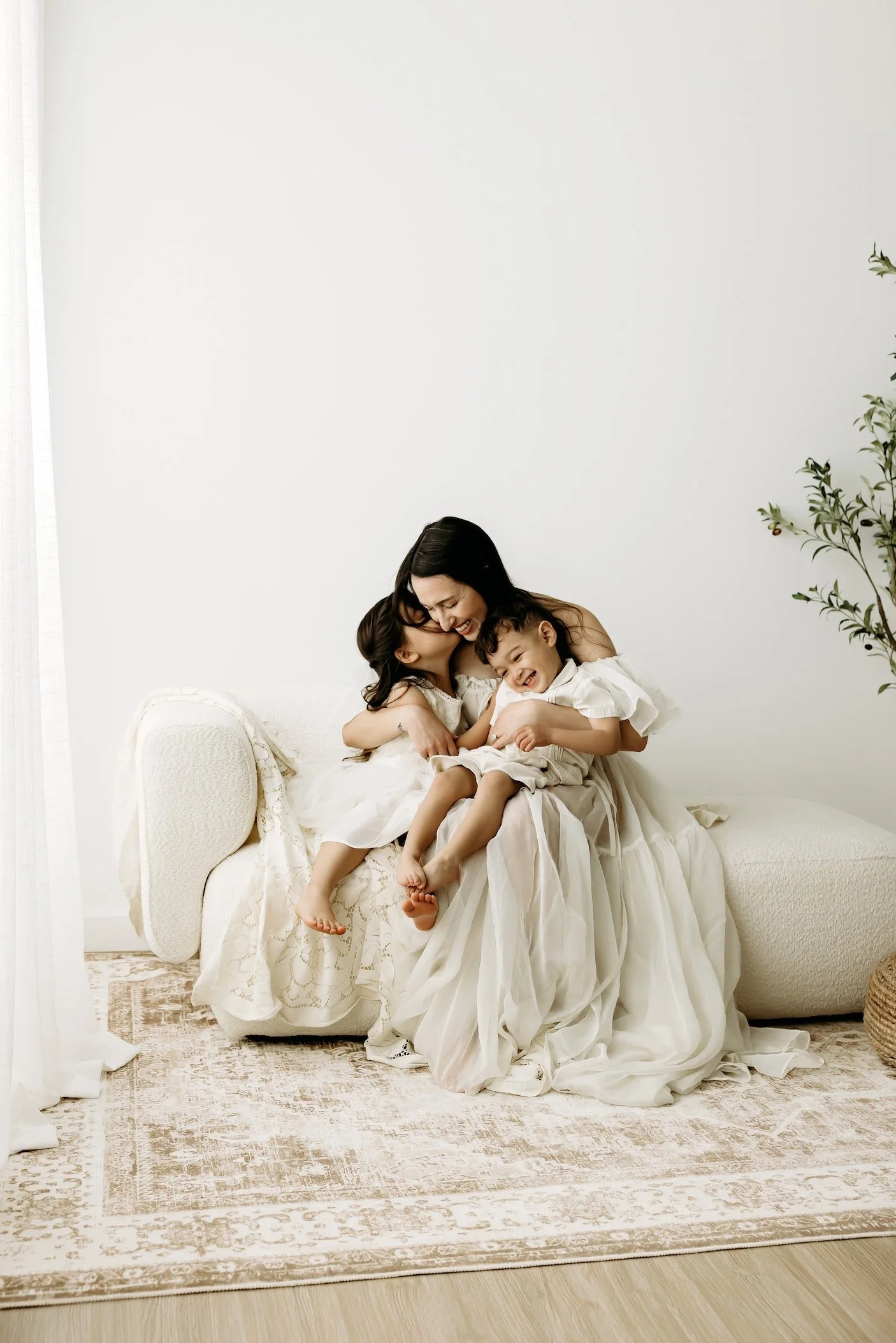 A woman and two children sitting on a cream-colored sofa in a bright room with white walls, sharing a joyful moment, all dressed in timeless white clothing and light and airy photos. 
