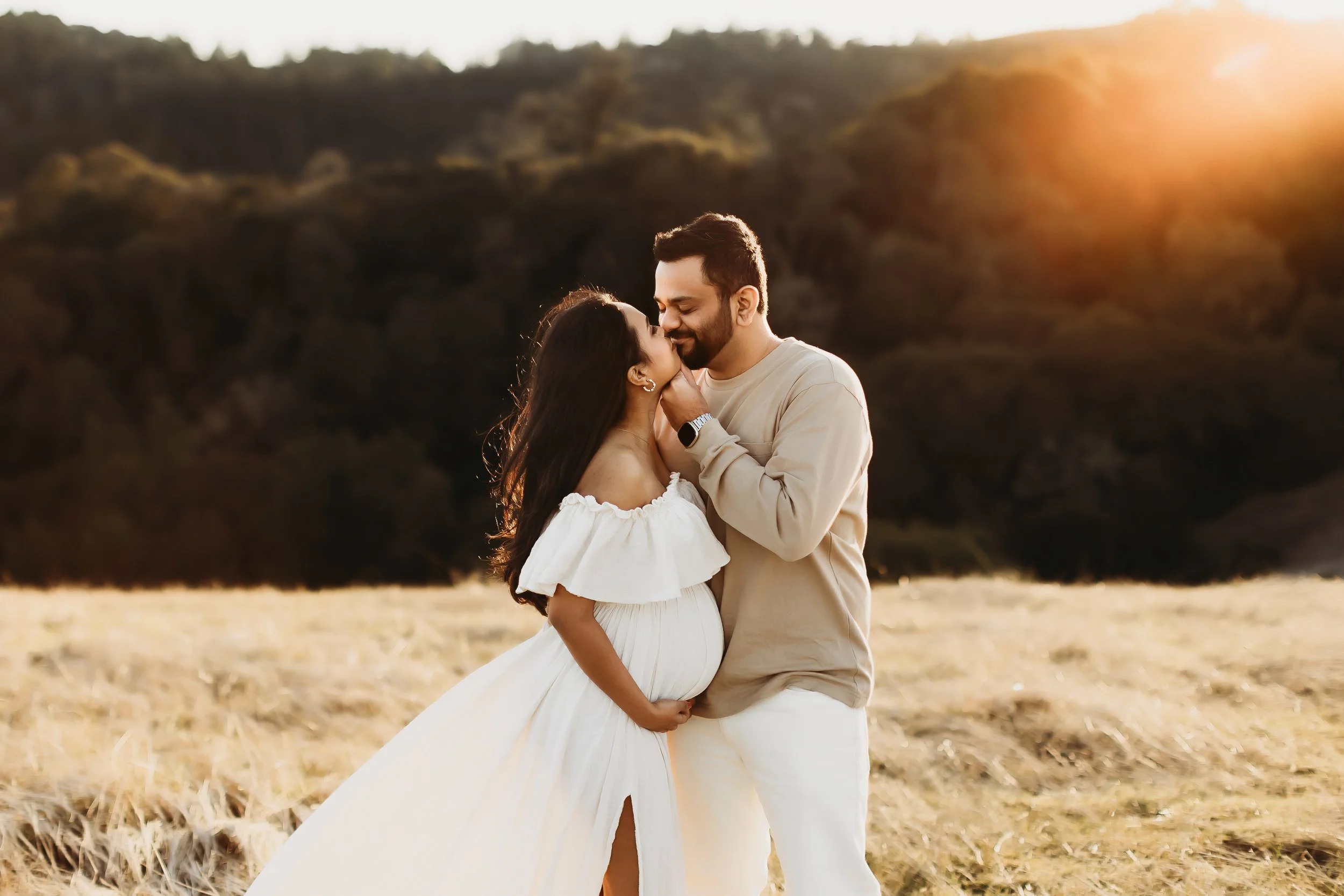 A couple hugging and kissing in a golden field during sunset, with rolling hills in the background.