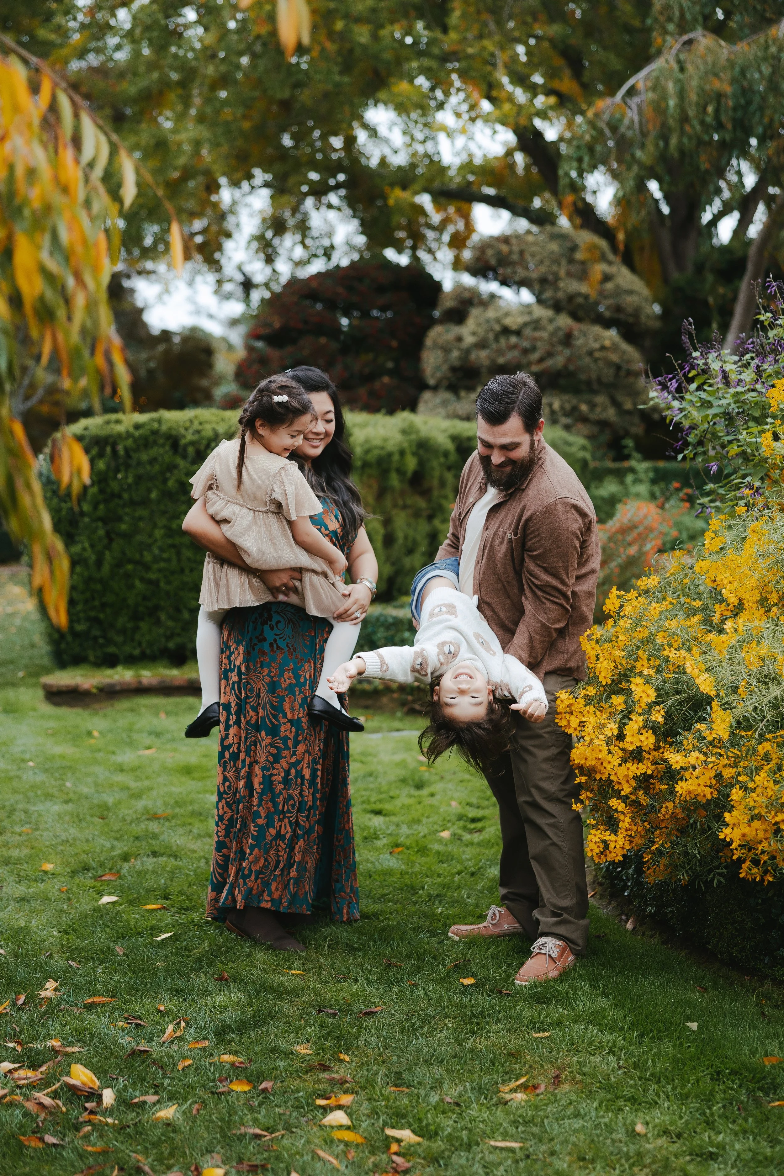 A family of four with a woman, a man, and two children, having fun outdoors in a lush garden with colorful flowers and trees. The man is holding one child upside down, and the woman is holding the other child, all smiling and enjoying the moment.