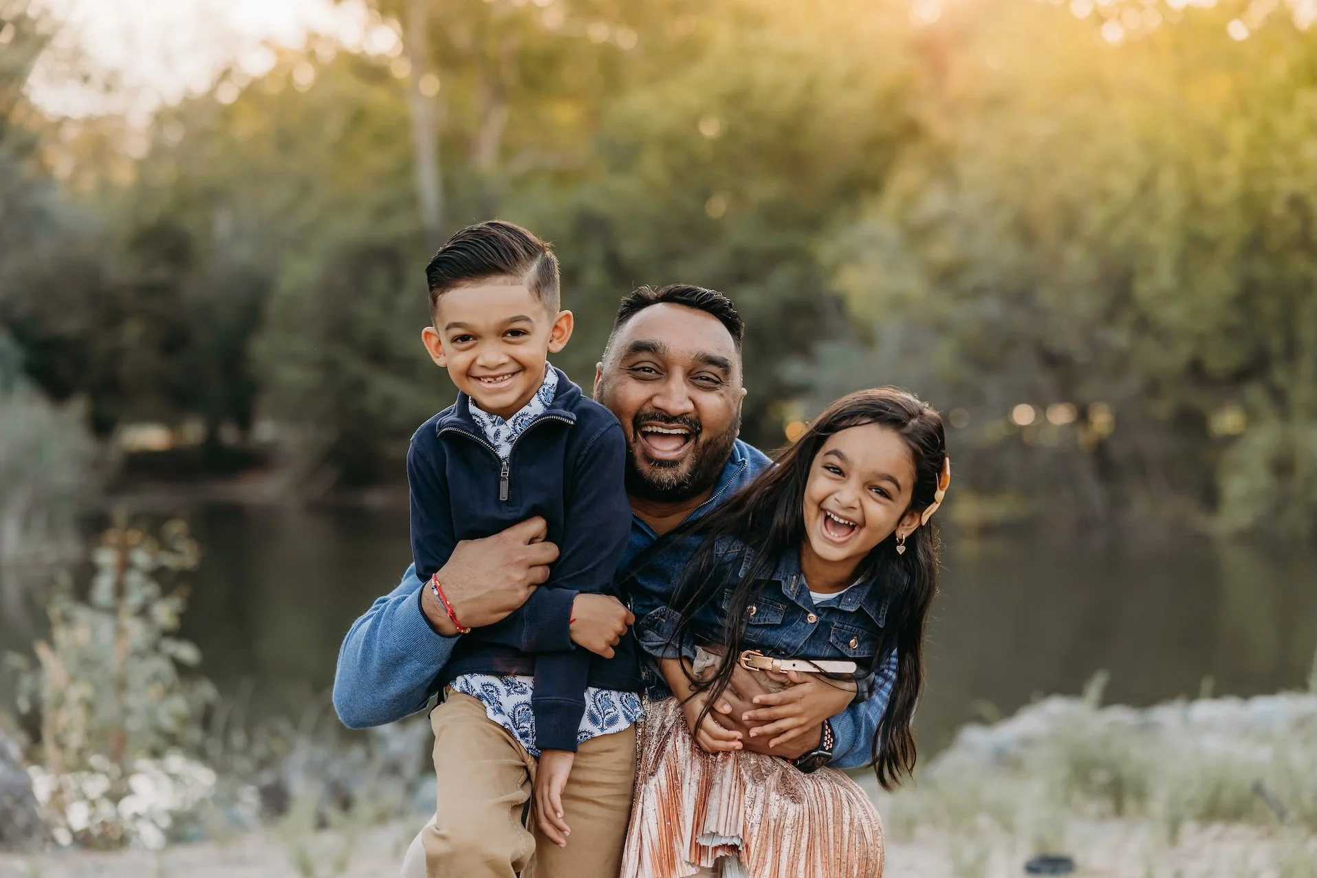 A smiling man with dark hair and beard holding a young boy on his shoulder, and a young girl standing beside them, outdoors near a body of water with trees in the background during sunset.