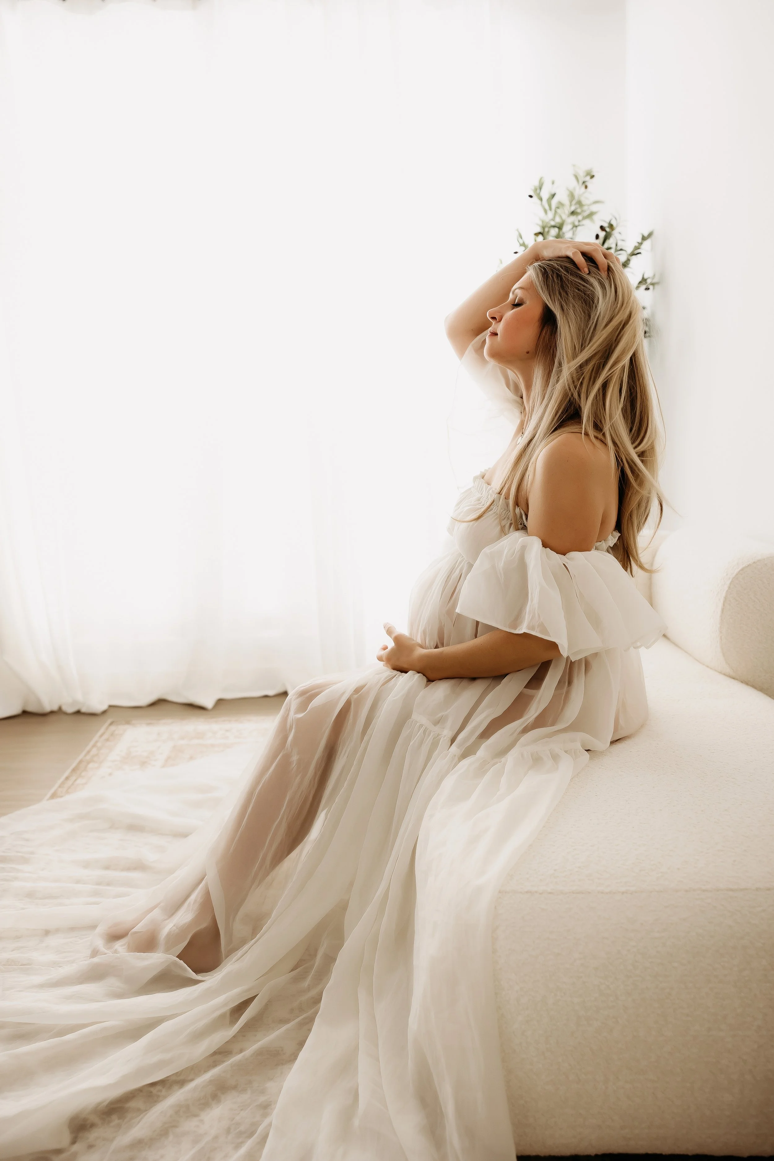 A woman with long blonde hair and closed eyes is sitting on a white couch, wearing a flowing white dress, with one hand on her head and the other resting on her lap, in a softly lit room with white walls and a potted plant in the background.