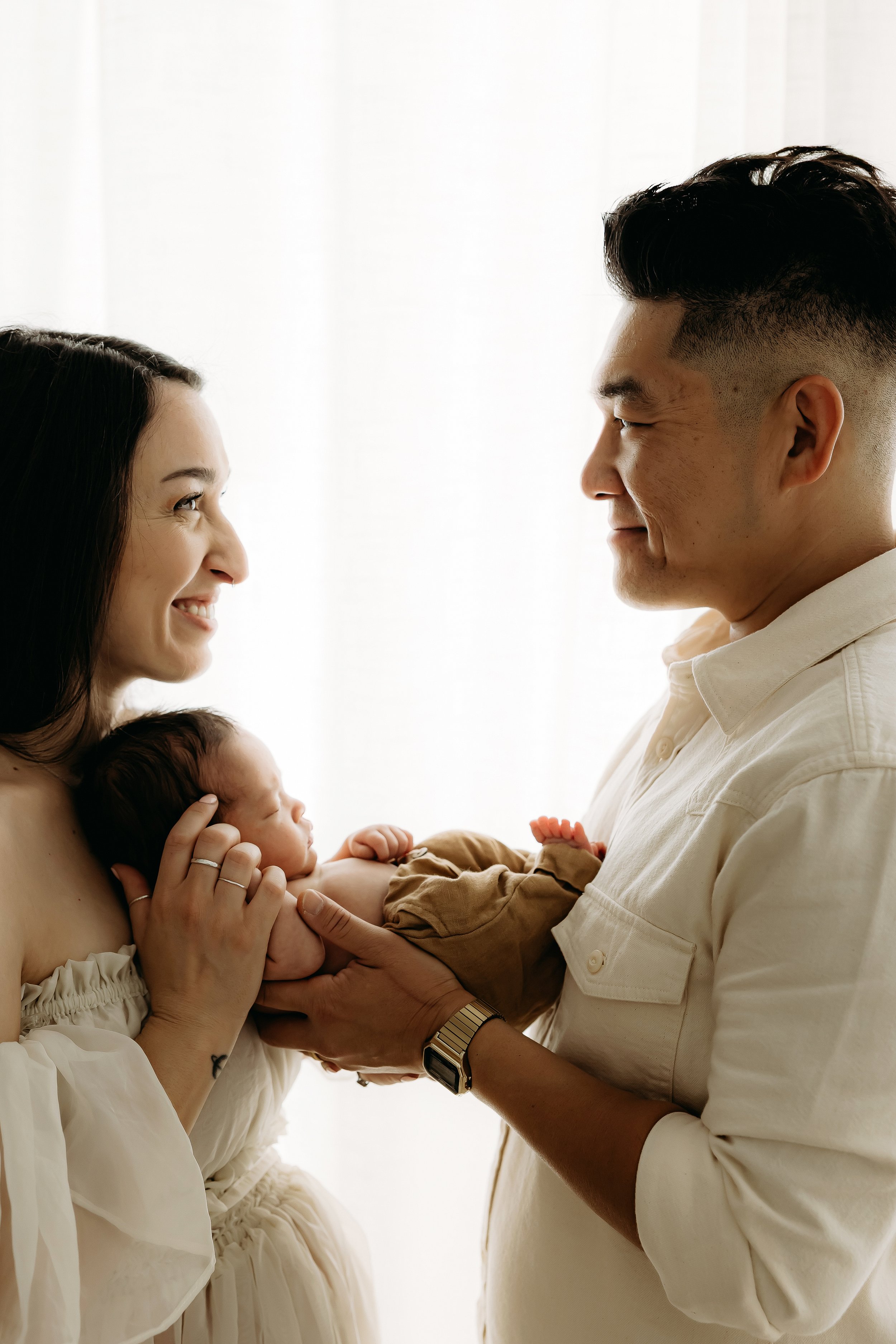 A woman and a man are holding a newborn baby. They are facing each other, smiling warmly. The woman has dark hair and is wearing a white blouse, while the man has dark, styled hair and is wearing a light-colored shirt. The baby is cradled in the woma