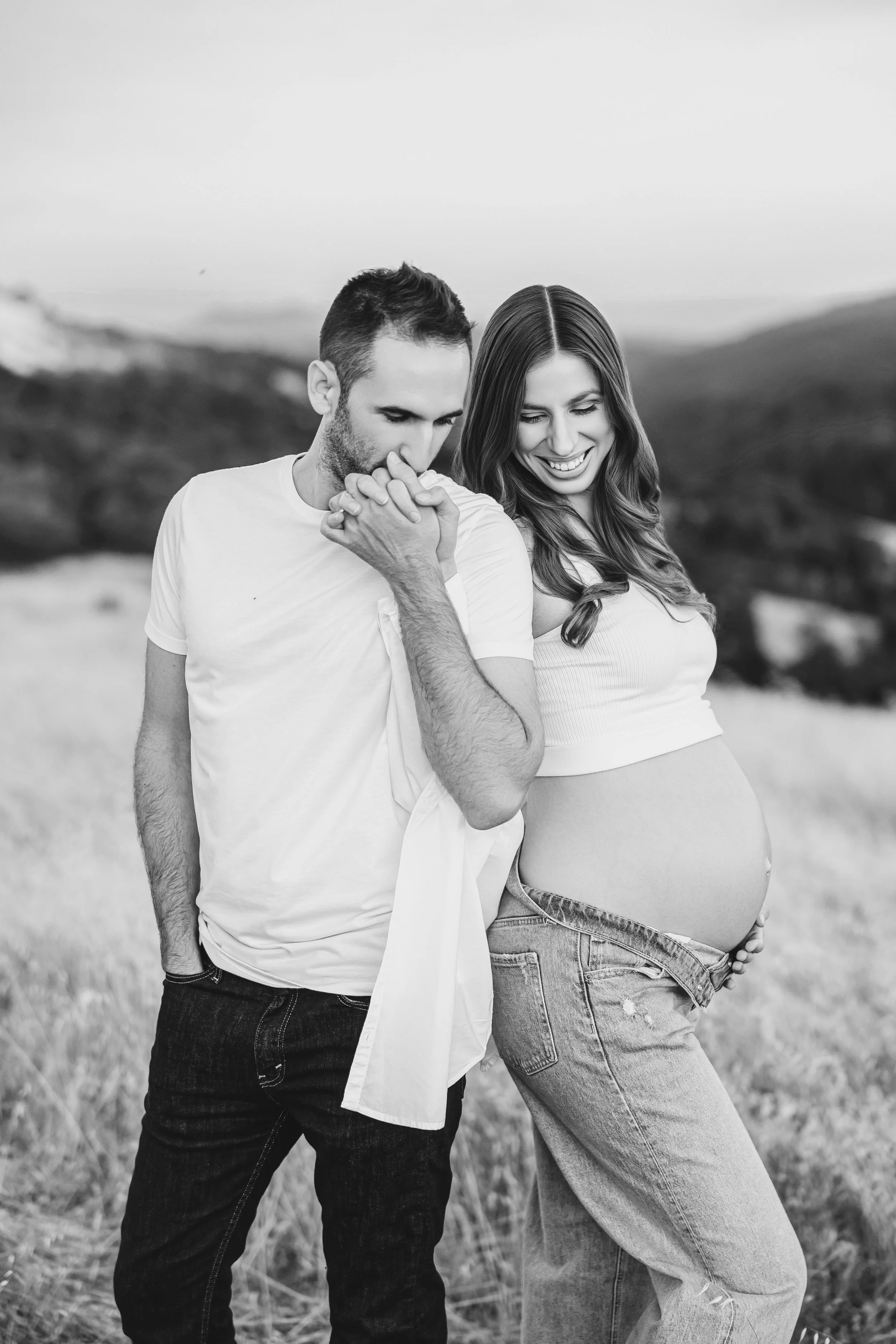 Black and white photo of a pregnant woman and a man outdoors, with the man whispering into the woman's ear and she smiling, standing in a grassy field with hills in the background.