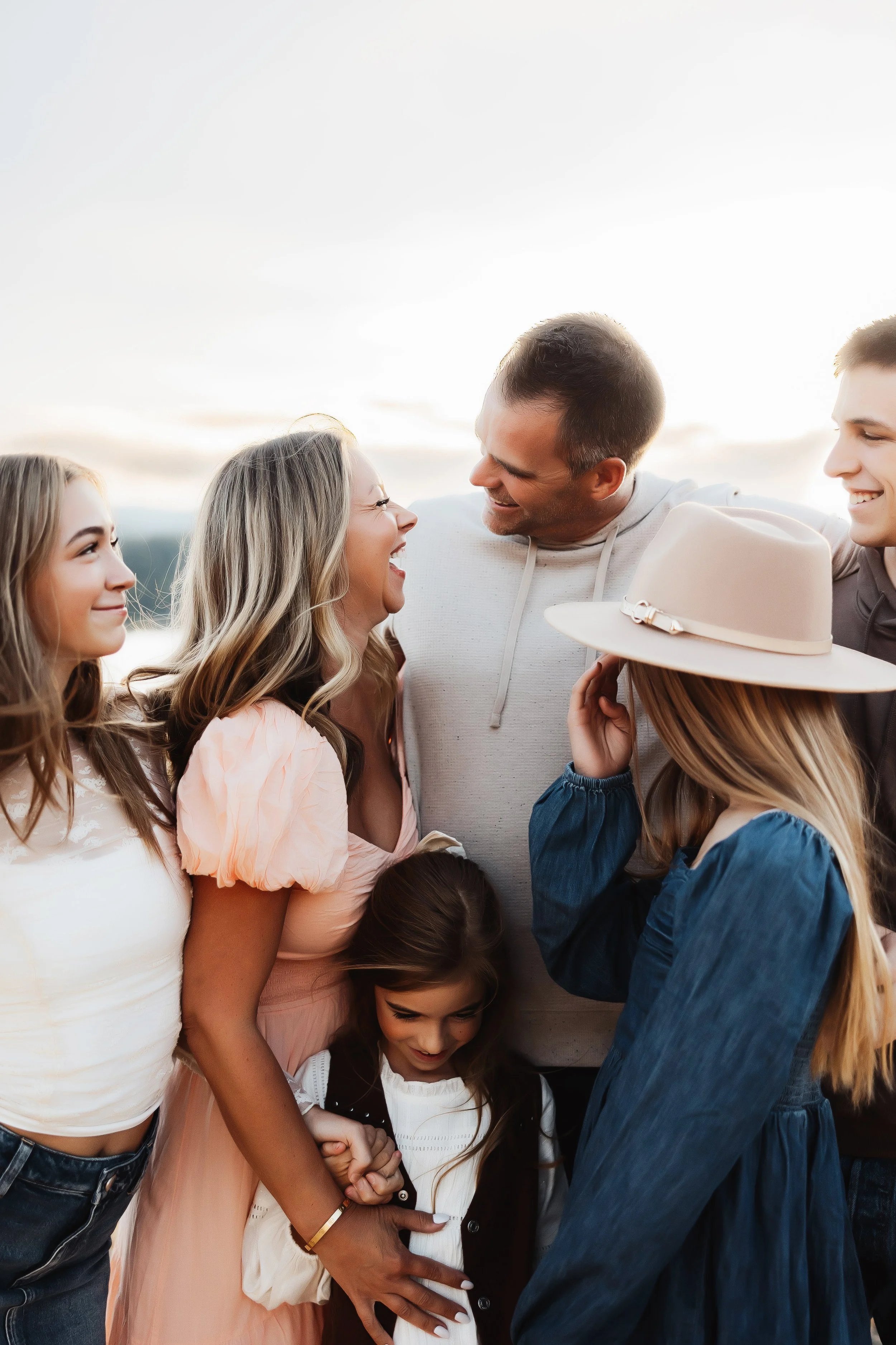 Group of six diverse women and men smiling, laughing, and embracing outdoors during sunset, wearing casual clothes.