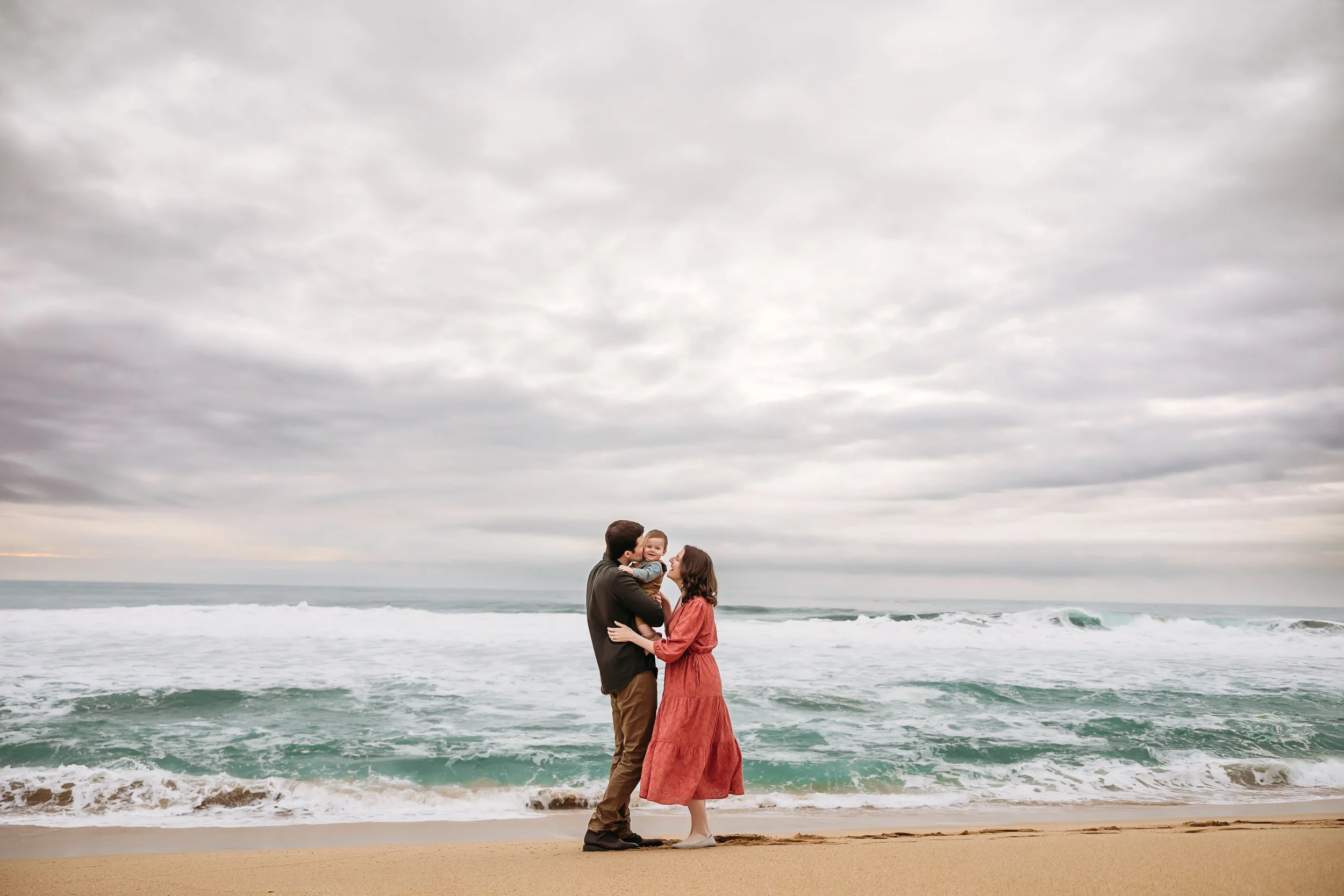 A family of three standing on the beach near the ocean, with two adults and a small child, embracing and smiling under a cloudy sky.