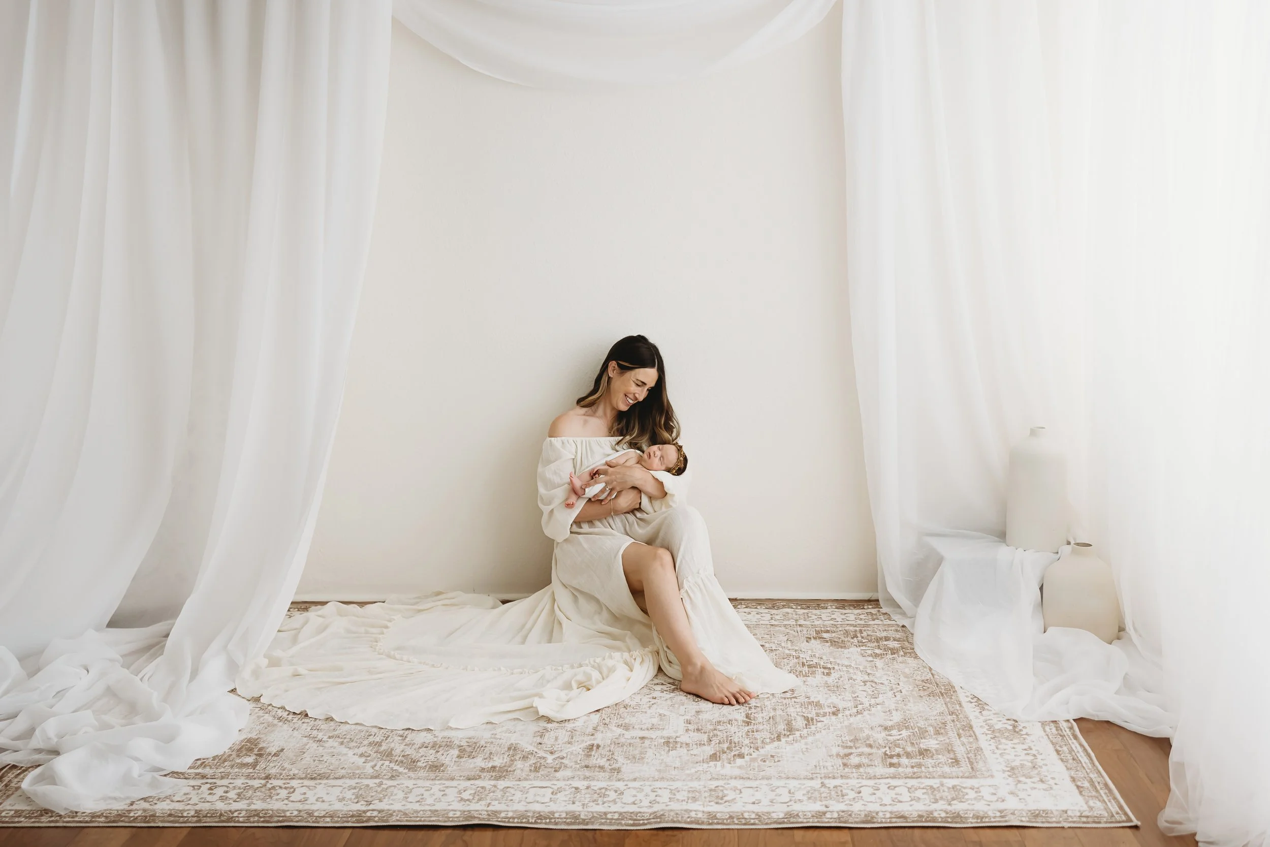 A woman with long brown hair, wearing a white off-shoulder dress, is sitting on a white sheet and holding a newborn baby in her arms. The setting is a minimalistic, bright room with white curtains hanging from the ceiling and a white wall behind them