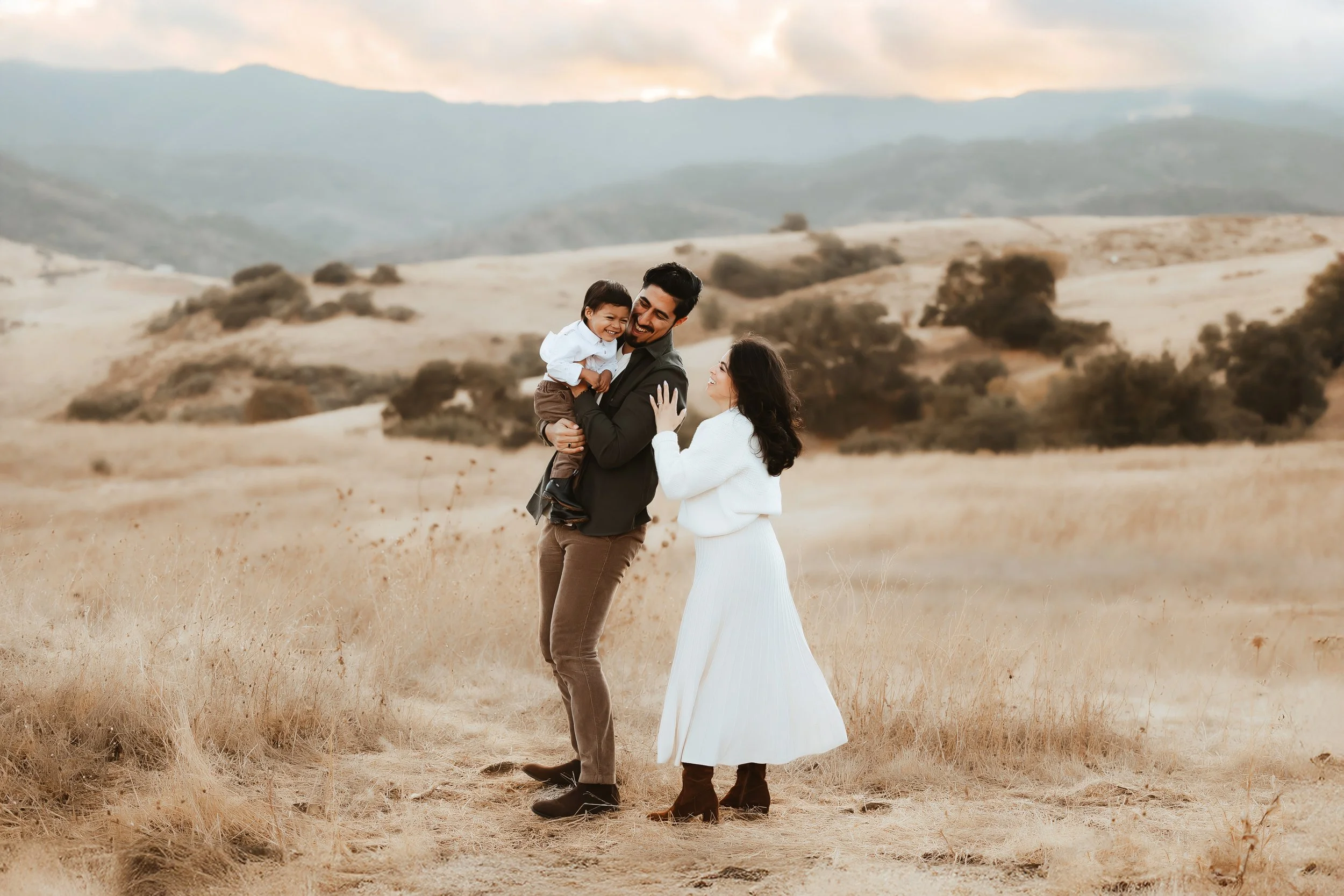 A man holding a child laughs while a woman in a white dress looks on in a field with dry grass and mountains in the background.