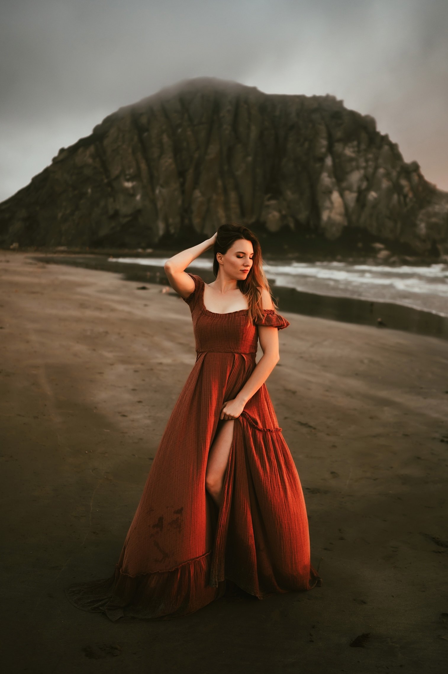 A woman in a rust-colored dress standing on a beach with a large, rocky mountain in the background. The sky is overcast and the woman is touching her hair with one hand, looking down slightly.