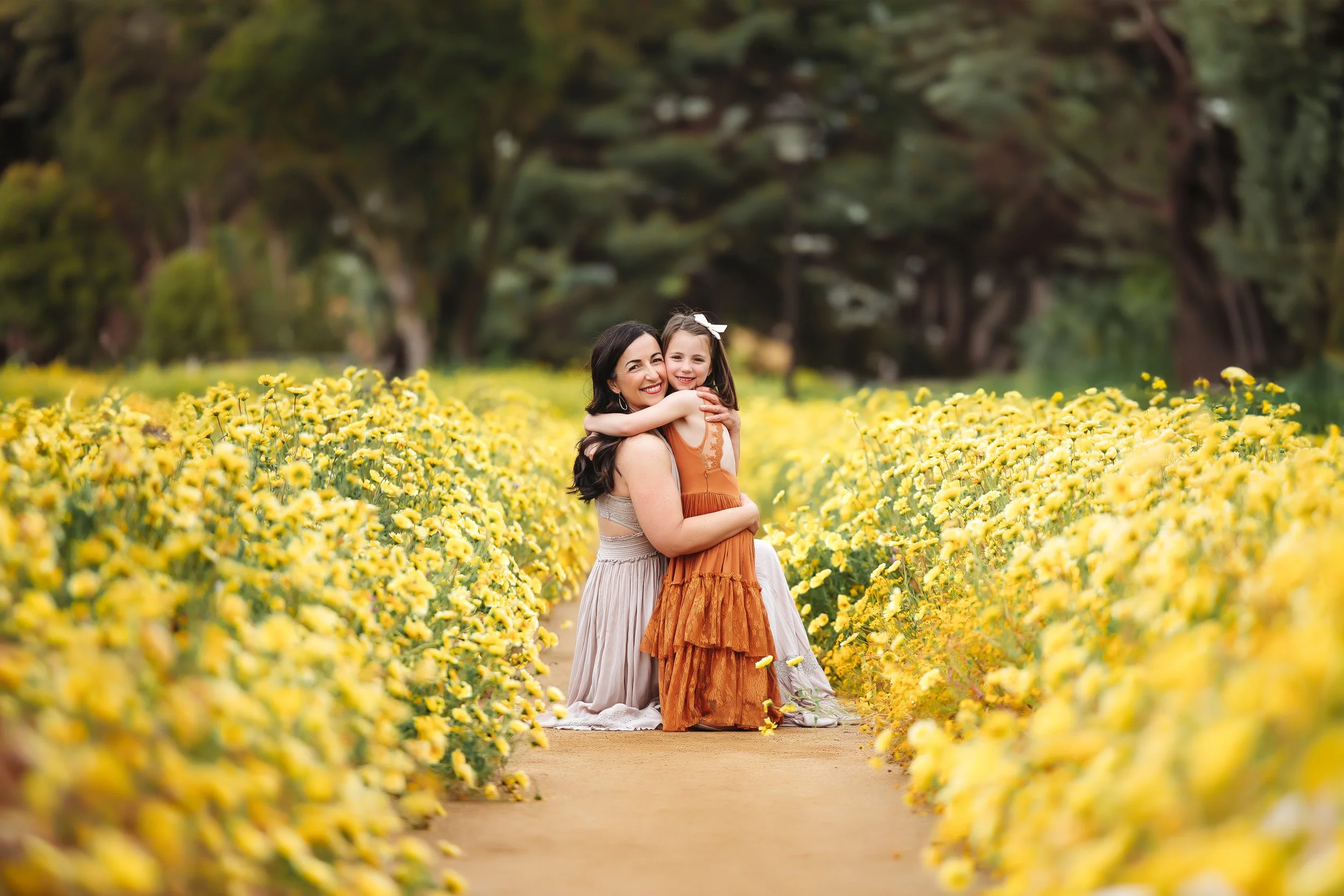 Mother and daughter hugging in a field of yellow flowers with trees in the background.