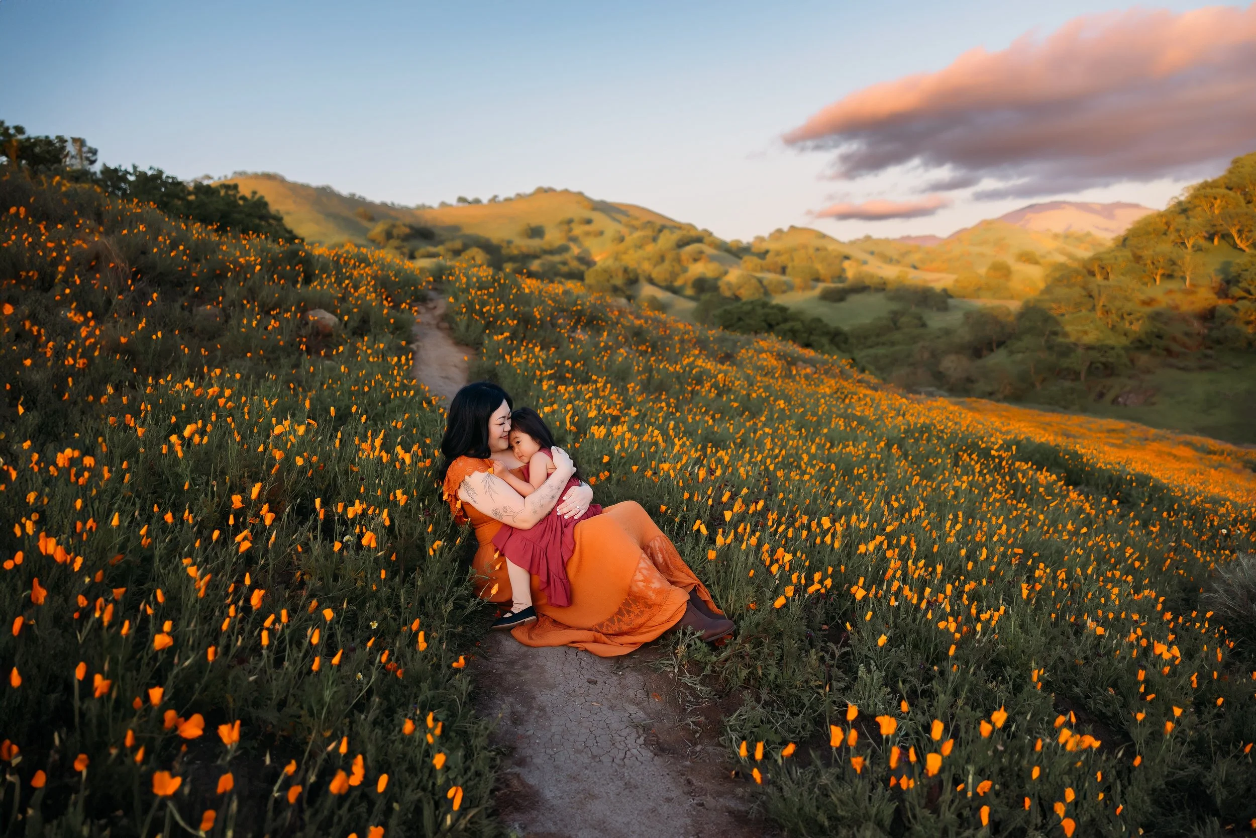 A woman and a girl sitting on a dirt trail in a field of orange poppies with rolling hills and trees in the background during sunset