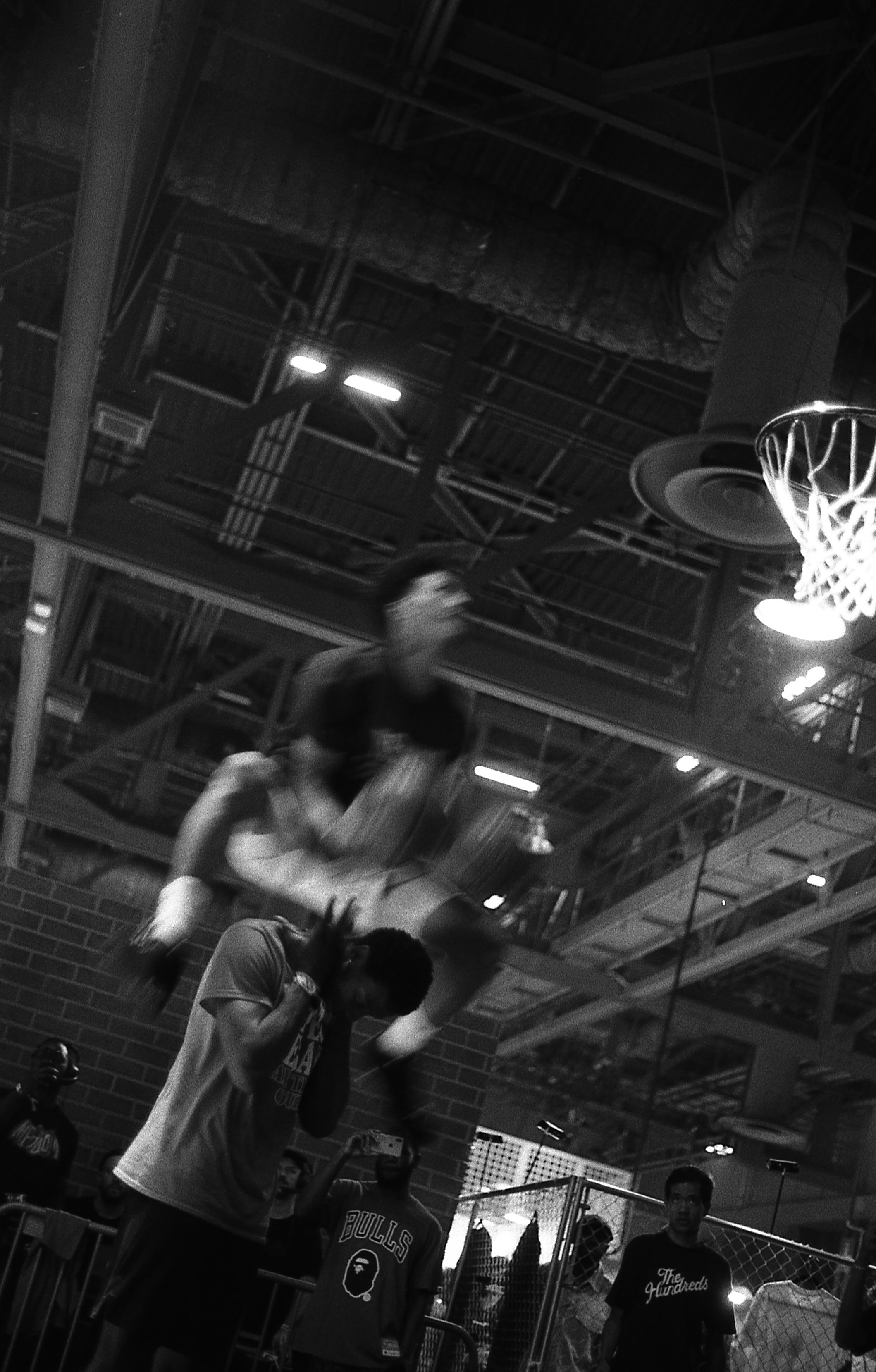 Two young men are playing basketball indoors. One is jumping and shooting while the other appears to be defending. Several people are watching nearby, with some taking photos. The setting is a gym with a high ceiling, exposed pipes, and a basketball 