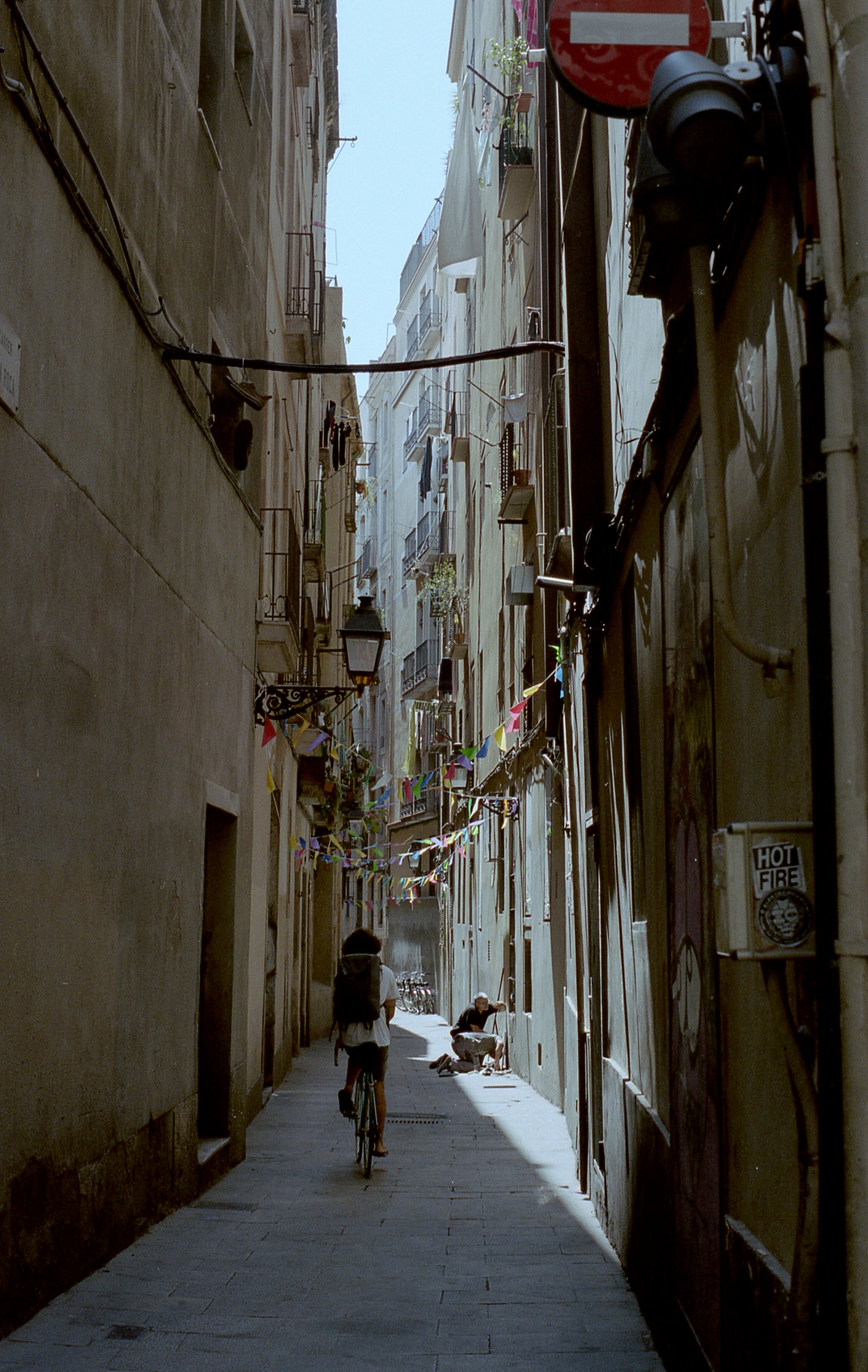 A narrow alleyway between tall buildings with stringed colorful flags hanging overhead. A person is riding a bicycle, while an older person is sitting on the ground, leaning against the wall. Various air conditioning units and street lamps are mounte
