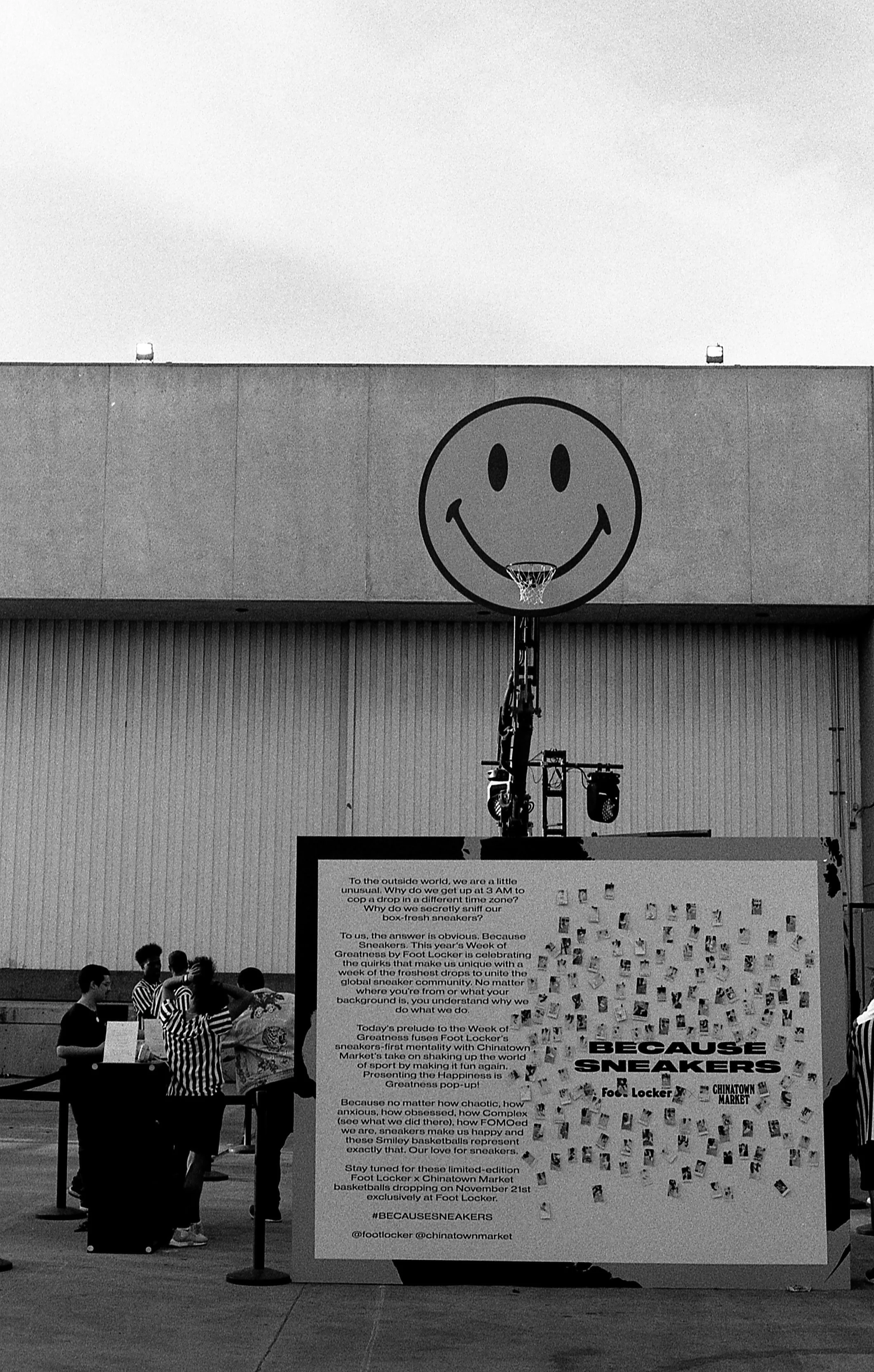 Black and white photo of a basketball hoop with a large smiley face at the backboard. To the left, there are a few people standing and talking.