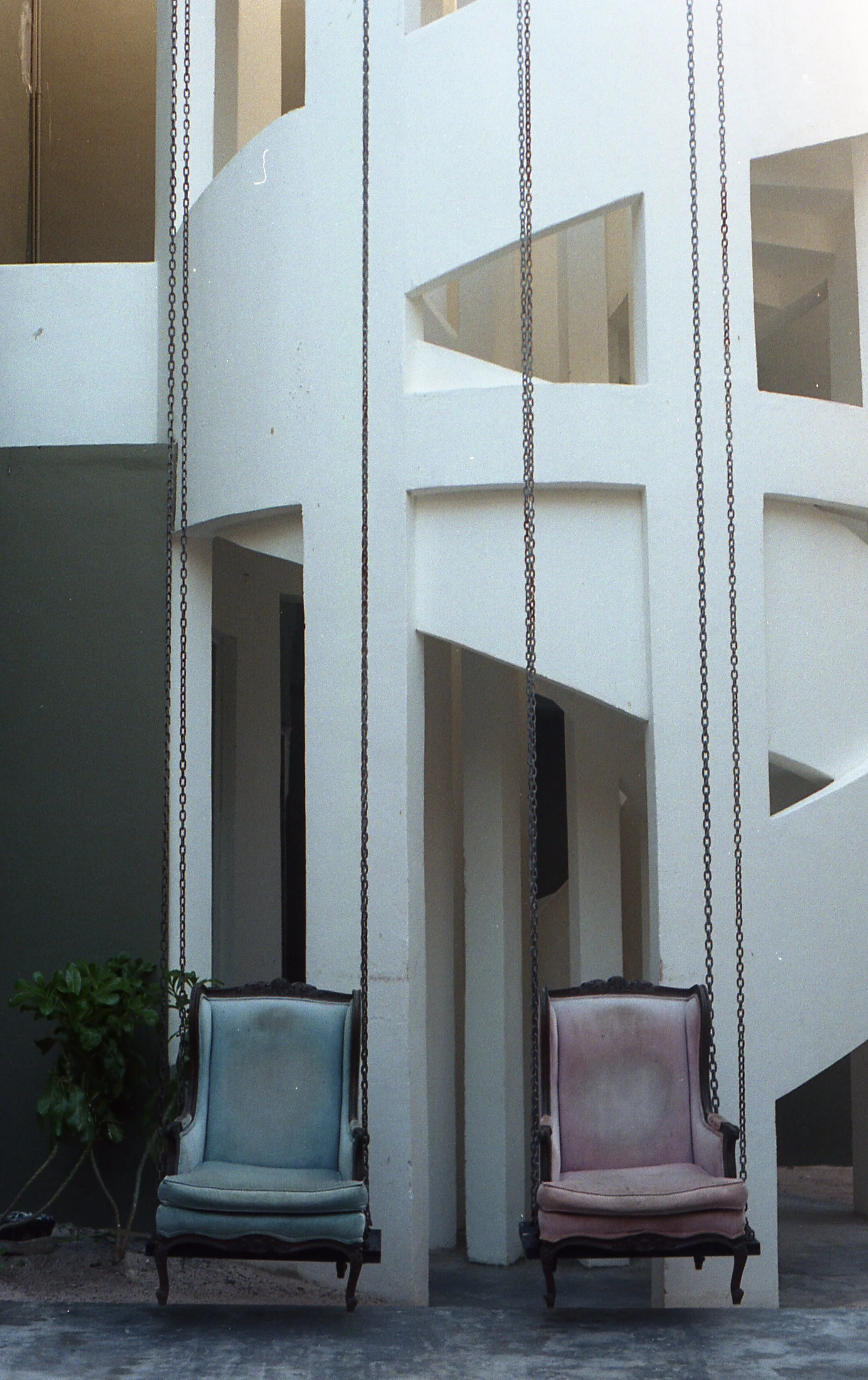 Two vintage-style armchairs, one blue and one pink, hanging on chains in front of a white staircase with geometric openings.