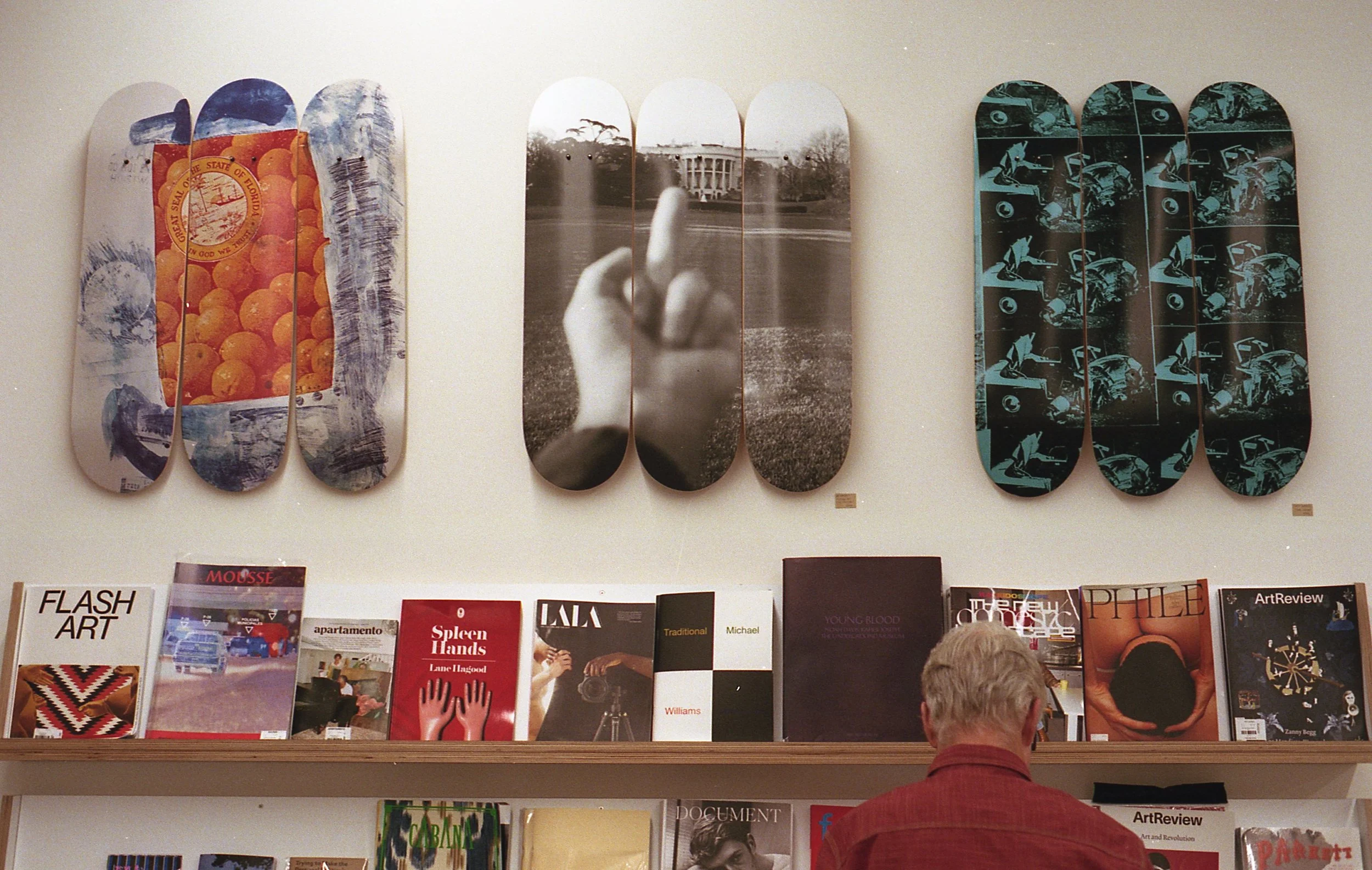 Gallery wall with three sets of painted skateboard decks and a magazine shelf below. The middle deck has a black-and-white photo of a hand giving a middle finger. The left deck features an image of oranges, and the right deck has a pattern of motorcy