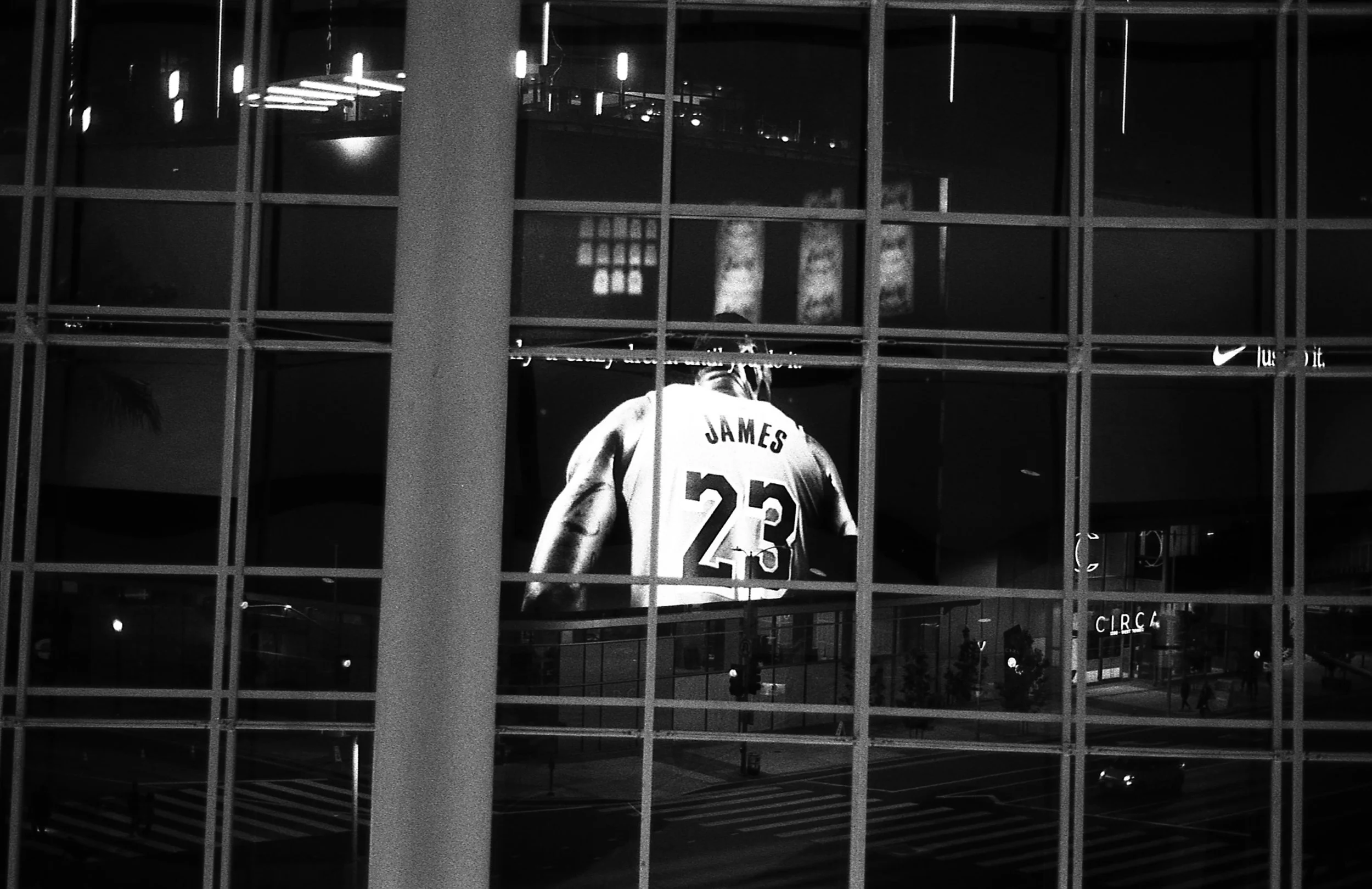 Large black-and-white photograph of a basketball player wearing a jersey with the name 'JAMES' and the number '23' displayed behind glass windows at night.