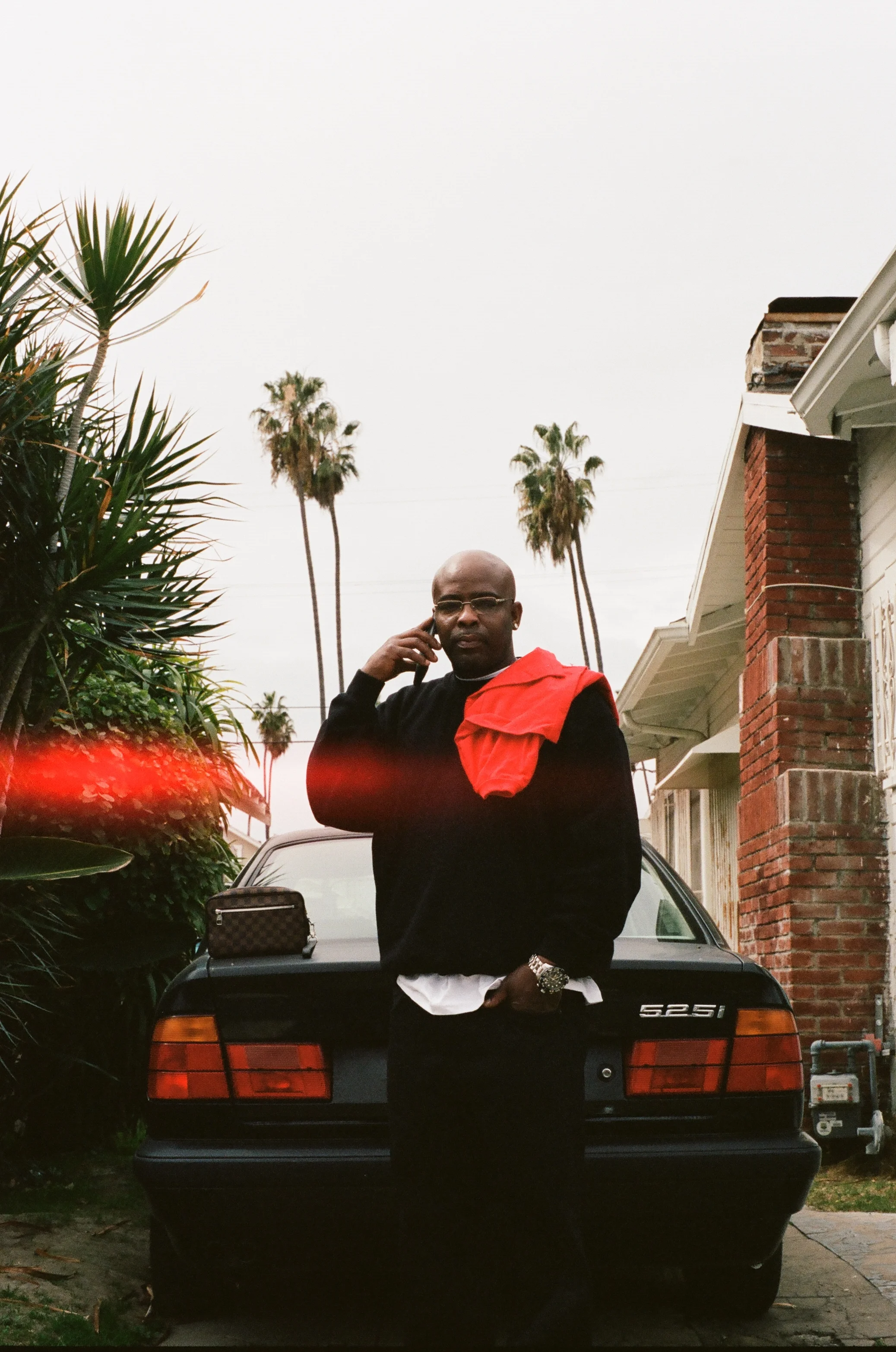 A man standing in front of a black Mercedes-Benz car, talking on a cellphone, with a house and tall palm trees in the background.