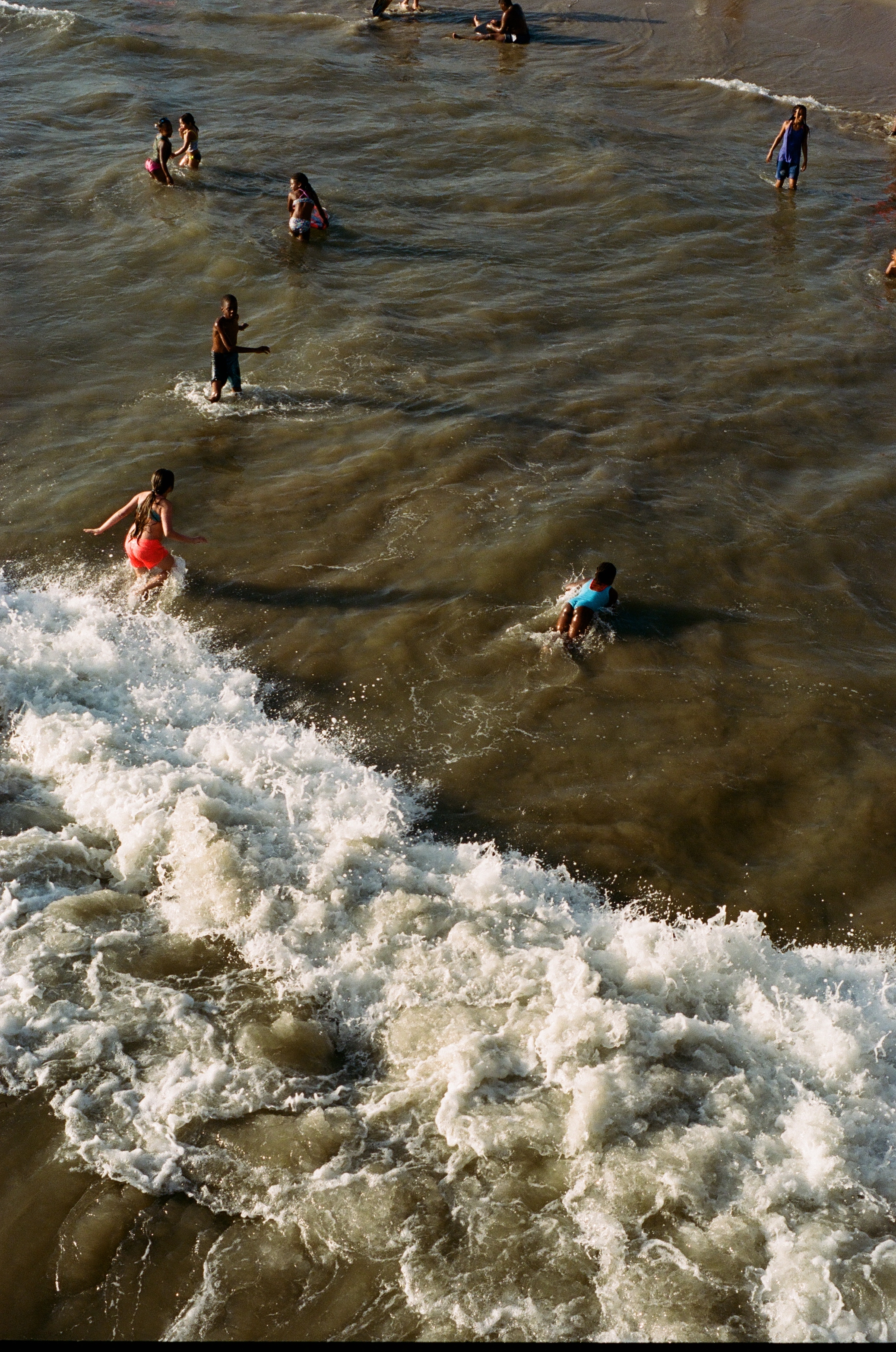 Children playing and swimming in the ocean, with some standing and others paddling or surfing, in a lively seaside scene.