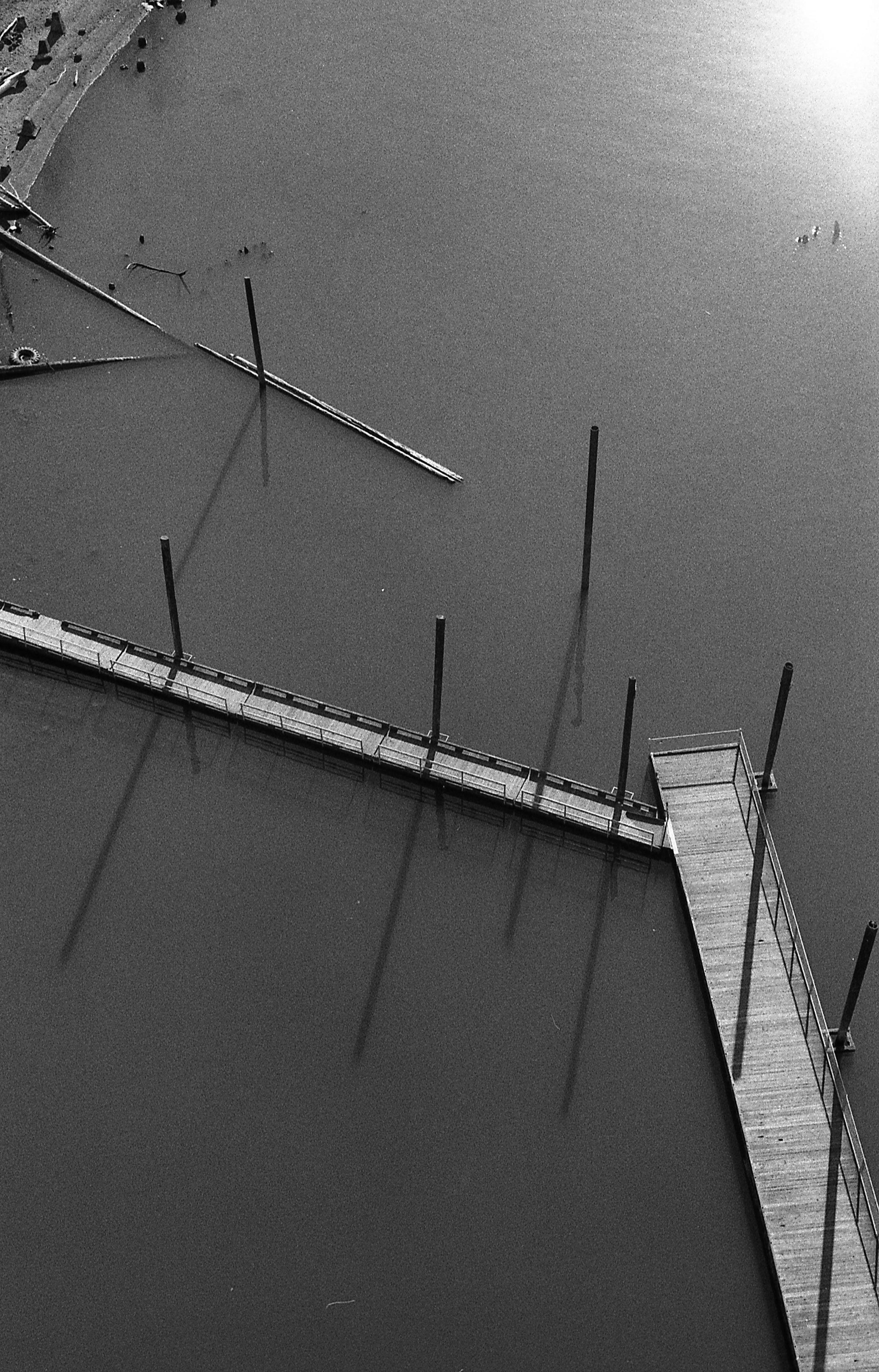 Black and white photo of a pier extending into a calm body of water, with reflections of the pier's posts visible in the water. There are a few scattered objects and debris on the water surface near the pier.