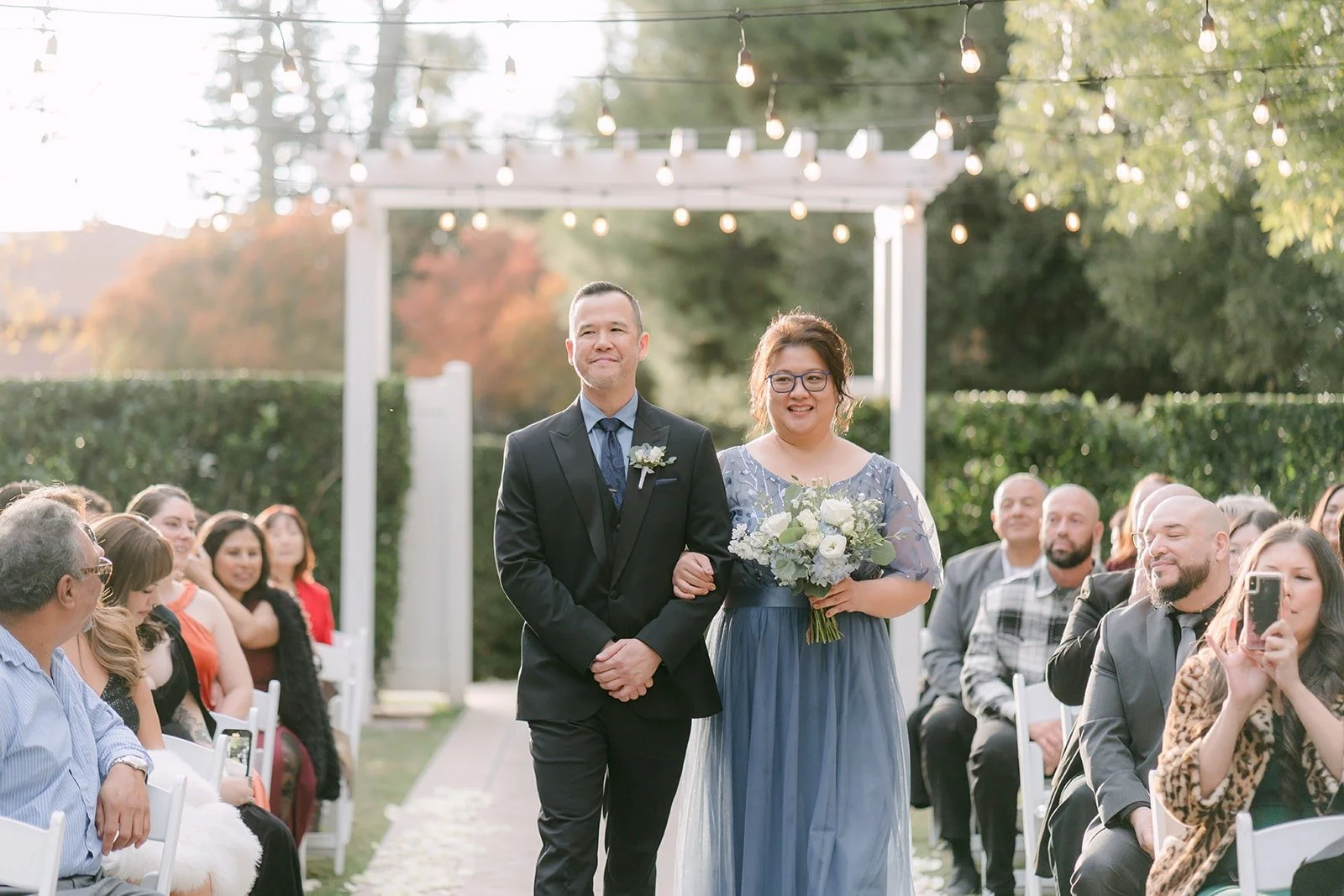 A groomsman and bridesmaid walk down the aisle under string lights