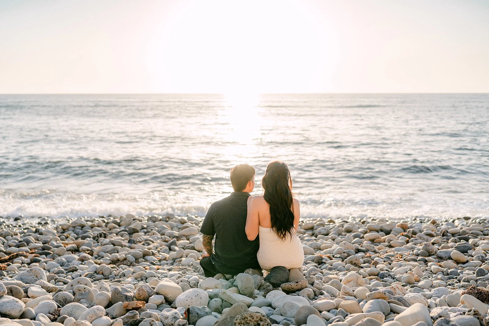 Victoria and Andrew sit side by side on the rocks facing the sunset.