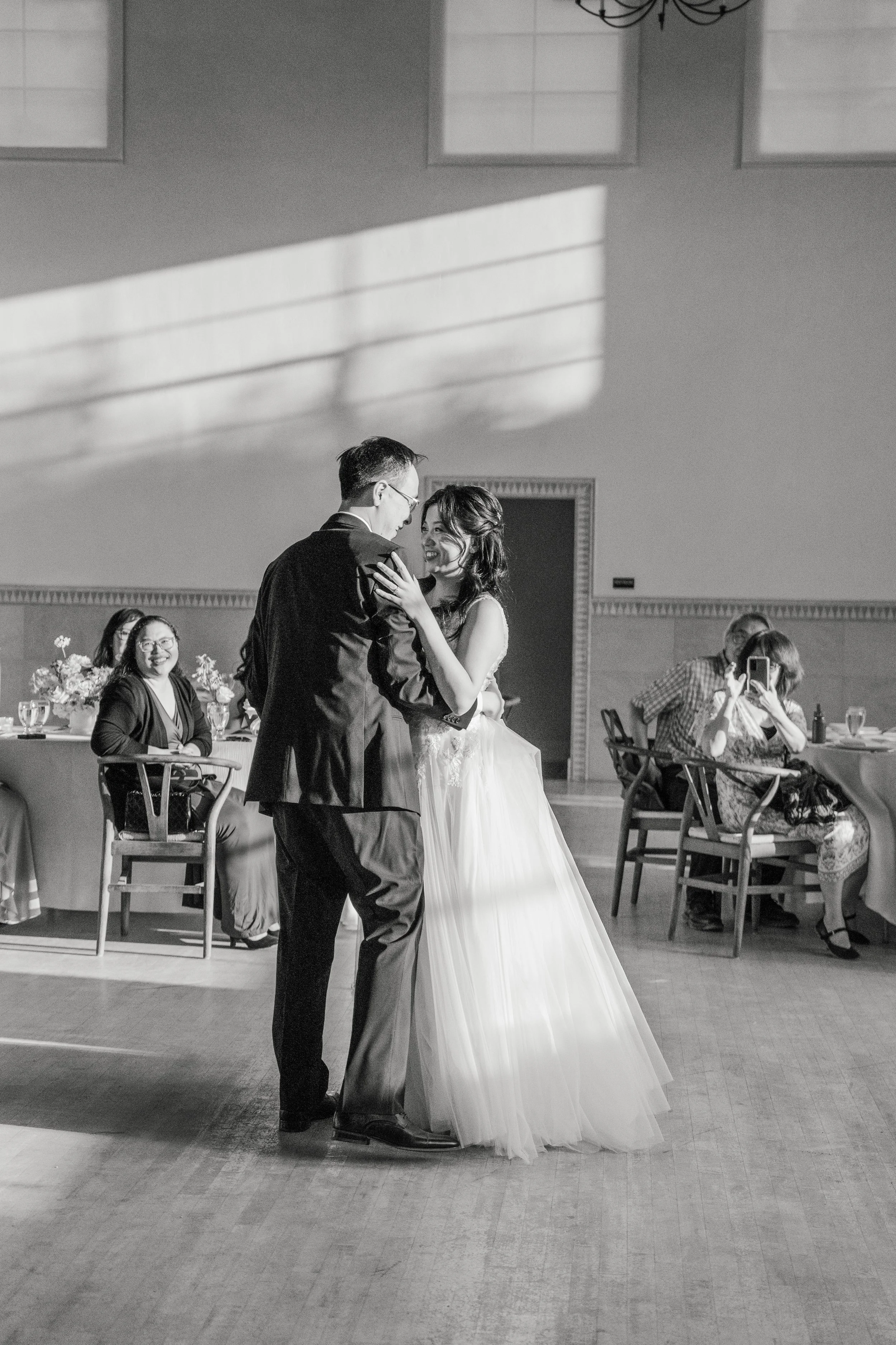 A bride and groom are dancing together at their wedding in a large hall, surrounded by seated guests and floral arrangements.
