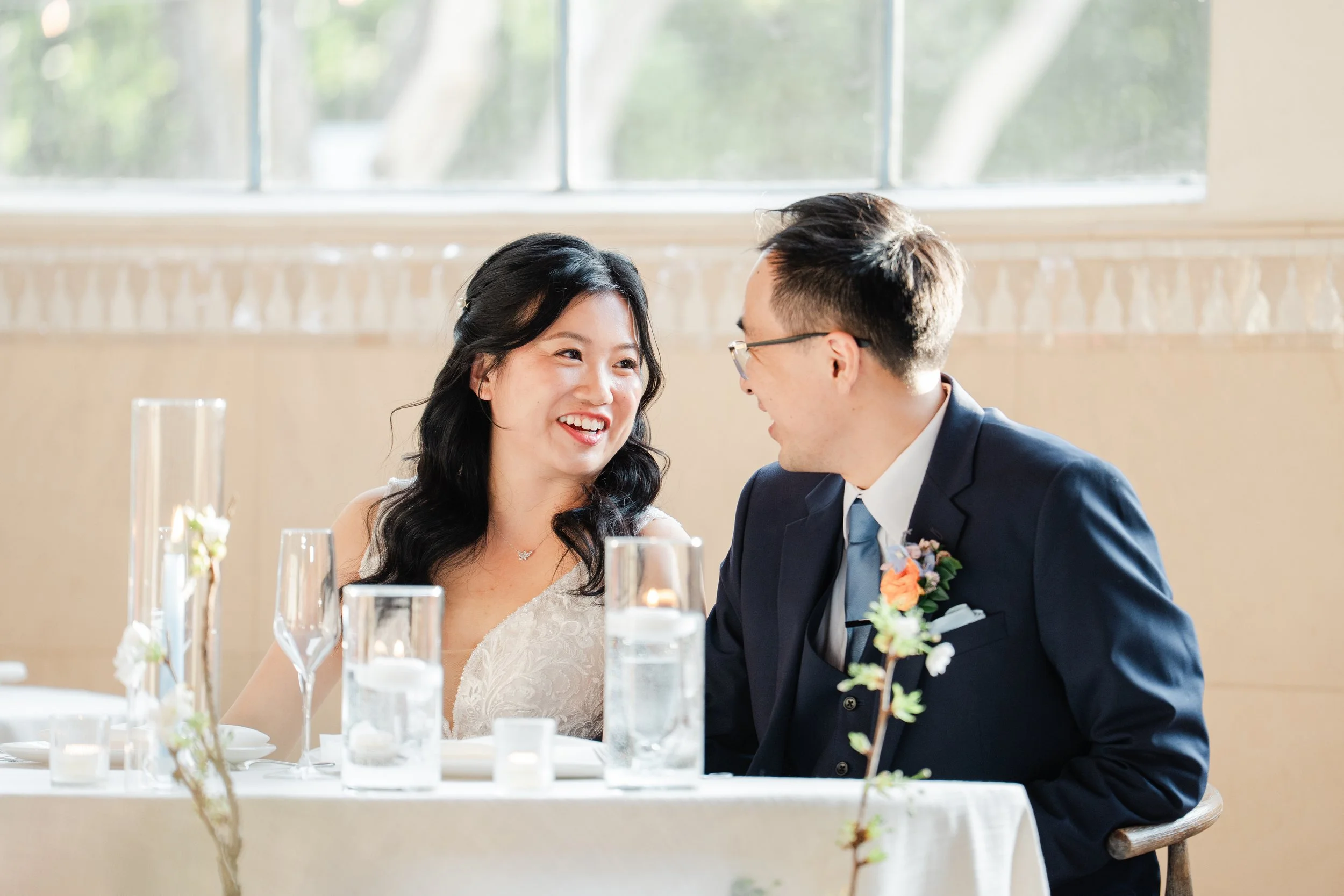 A smiling woman and man in formal attire sitting close at a table during a wedding reception. The woman is wearing a white lace dress, and the man is in a dark suit with a blue tie and boutonniere. There are glass and floral decorations on the table.