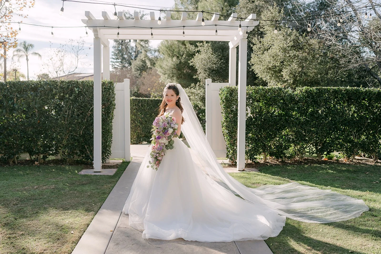 Kate under the white pergola with her veil spread across the aisle.