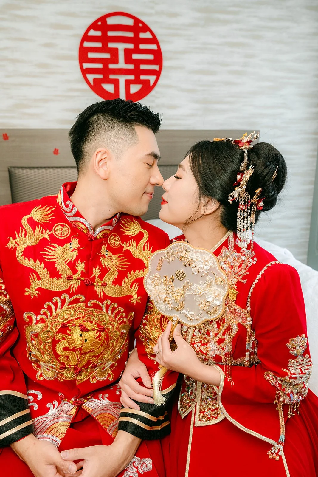 A groom kissing the bride's forehead as she holds a fan in front of a double happiness sign.